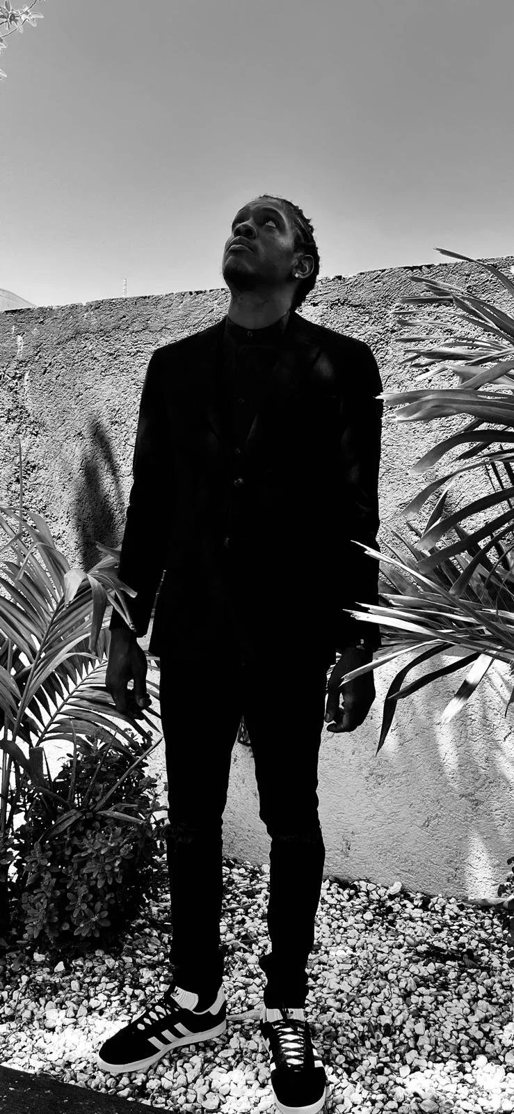 A man standing outdoors against a textured wall, wearing a dark suit and sneakers, looking upward in a black-and-white photo.
