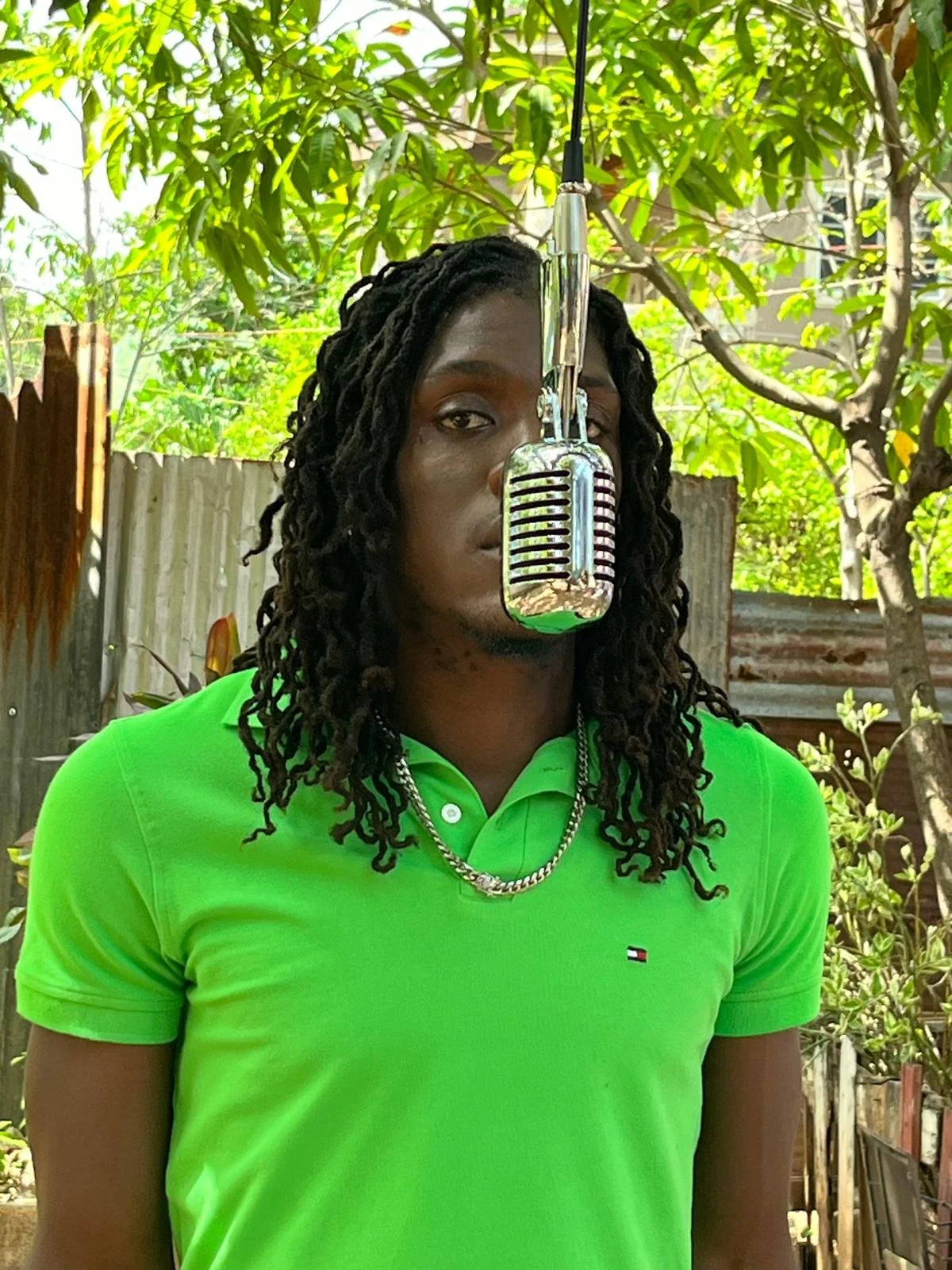 A man with dreadlocks is standing outdoors, wearing a bright green polo shirt, a silver chain necklace, and is partially obscured by a vintage-style microphone hanging in front of his face. Behind him, there are trees and a metal fence.