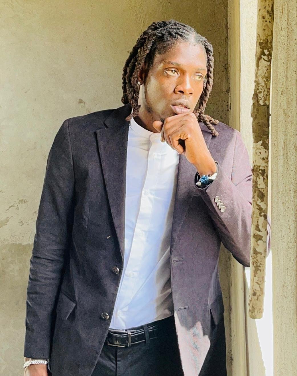 A man with dreadlocks in a suit and white shirt, standing near a window, thoughtfully posing with one hand on his chin.