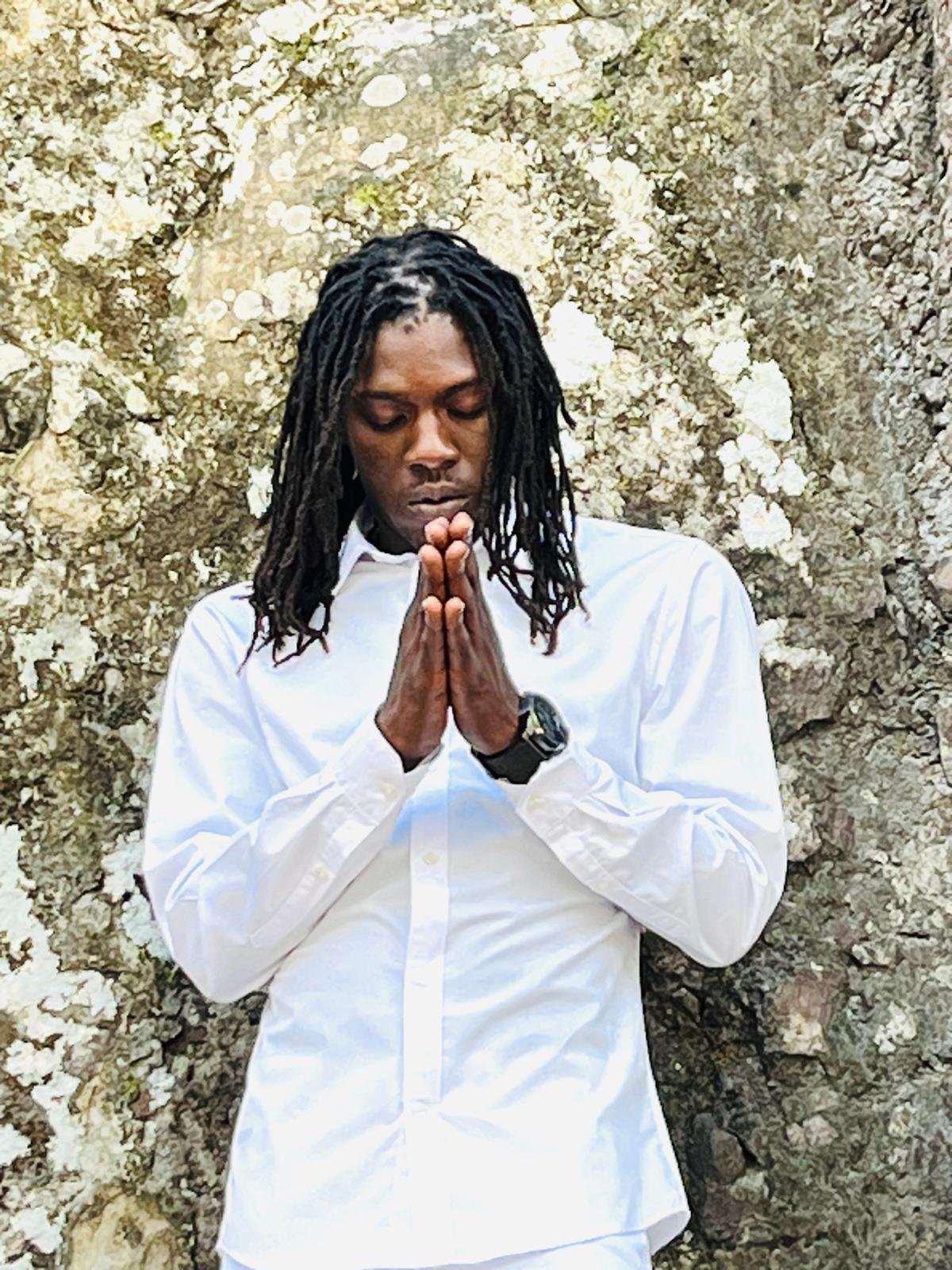 A man with dreadlocks wearing a white dress shirt and a black watch, standing with hands pressed together in a prayer pose in front of a rocky, textured background.