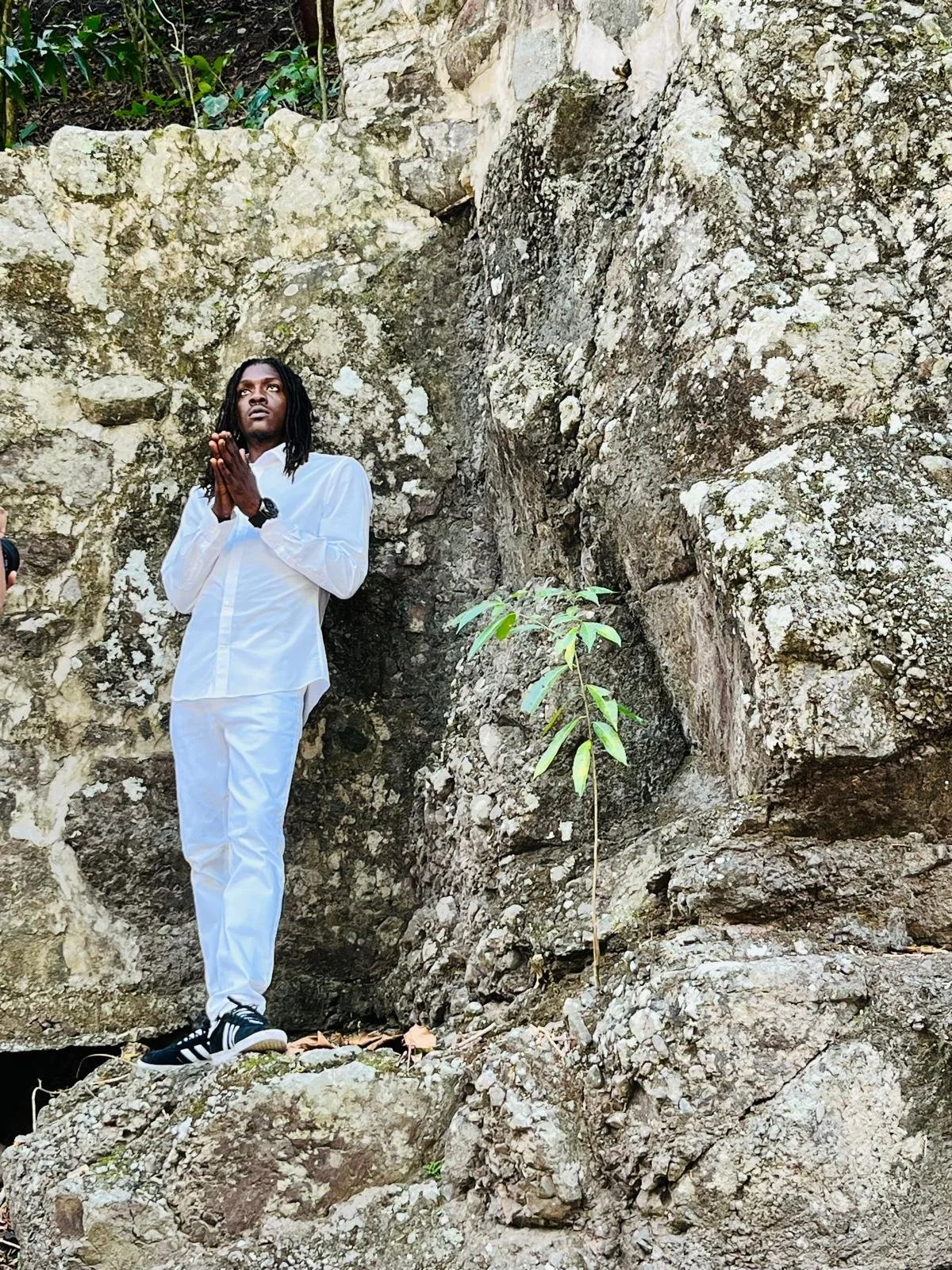 A man dressed in white pants and a white shirt standing on large rocks in a natural setting with a rocky cliff and greenery in the background, holding his hands together in a prayer-like gesture.