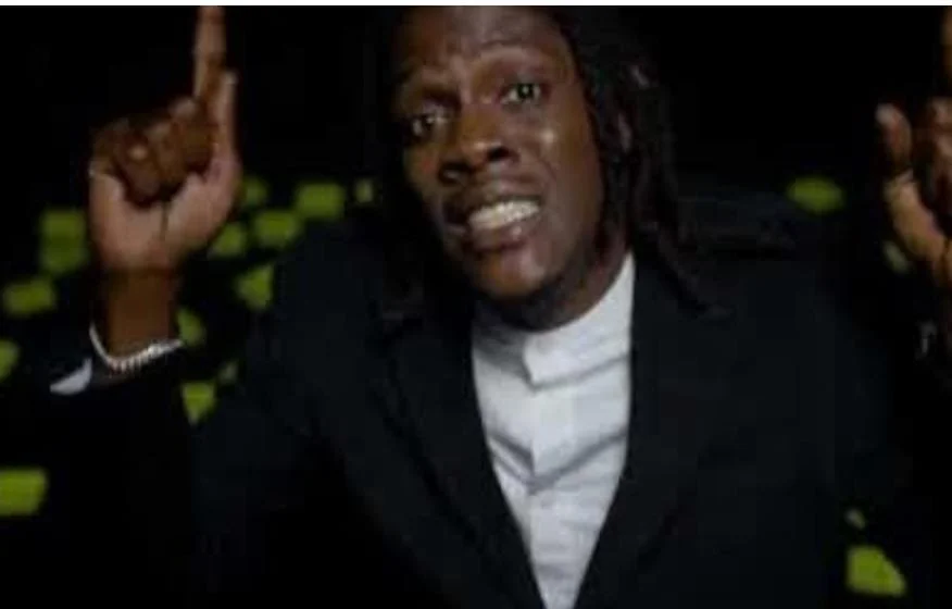 A man with braided hair wearing a black suit and white shirt, gesturing with his right index finger, speaking or singing passionately.