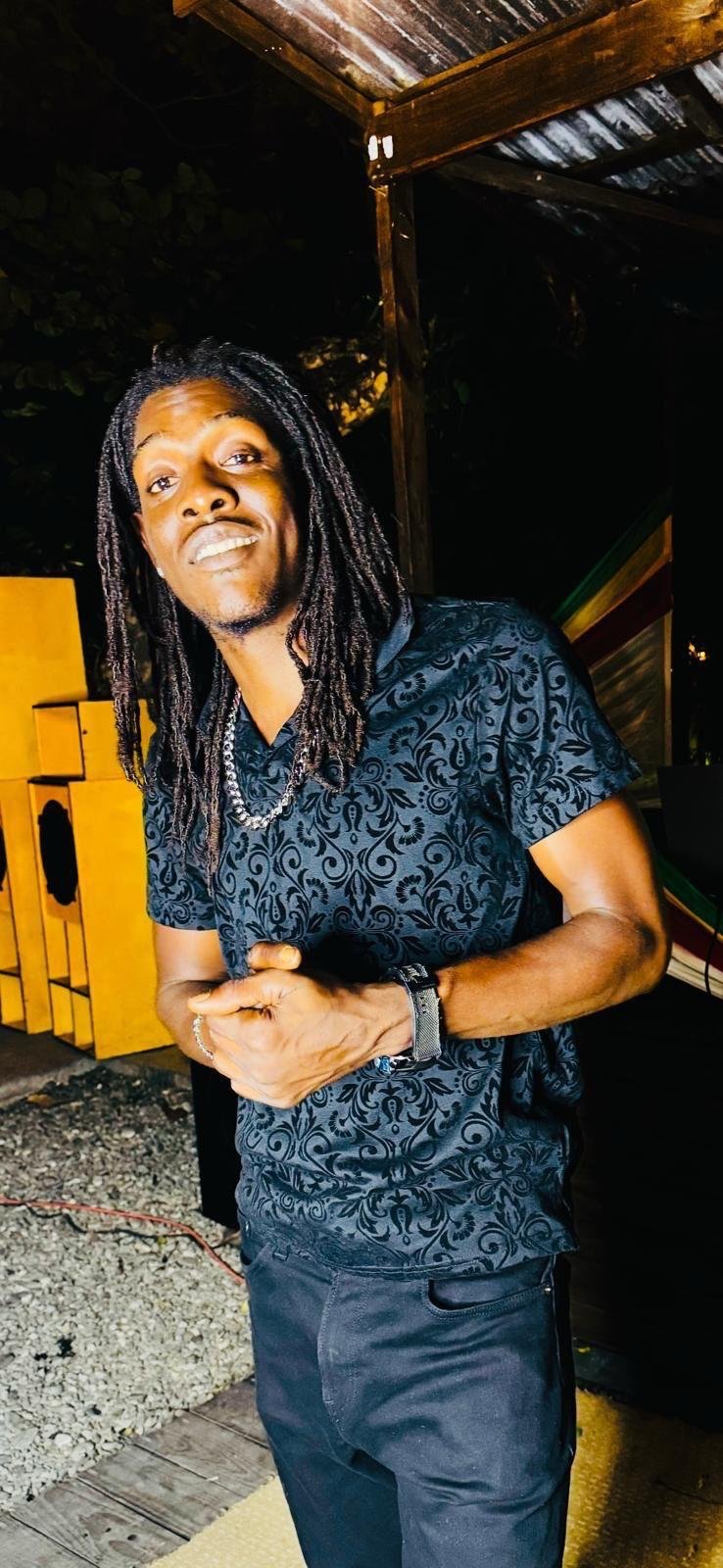 A man with long dreadlocks and a mustache wearing a black patterned short-sleeve shirt, a silver chain, and a wristwatch standing outdoors at night. He is smiling slightly and has his hands clasped together.