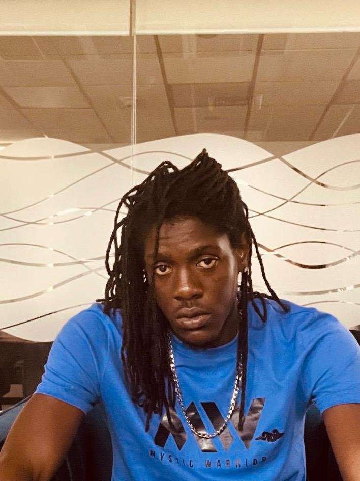 A young man with long dreadlocks wearing a blue t-shirt and a silver chain necklace, sitting indoors with a patterned wall behind him.