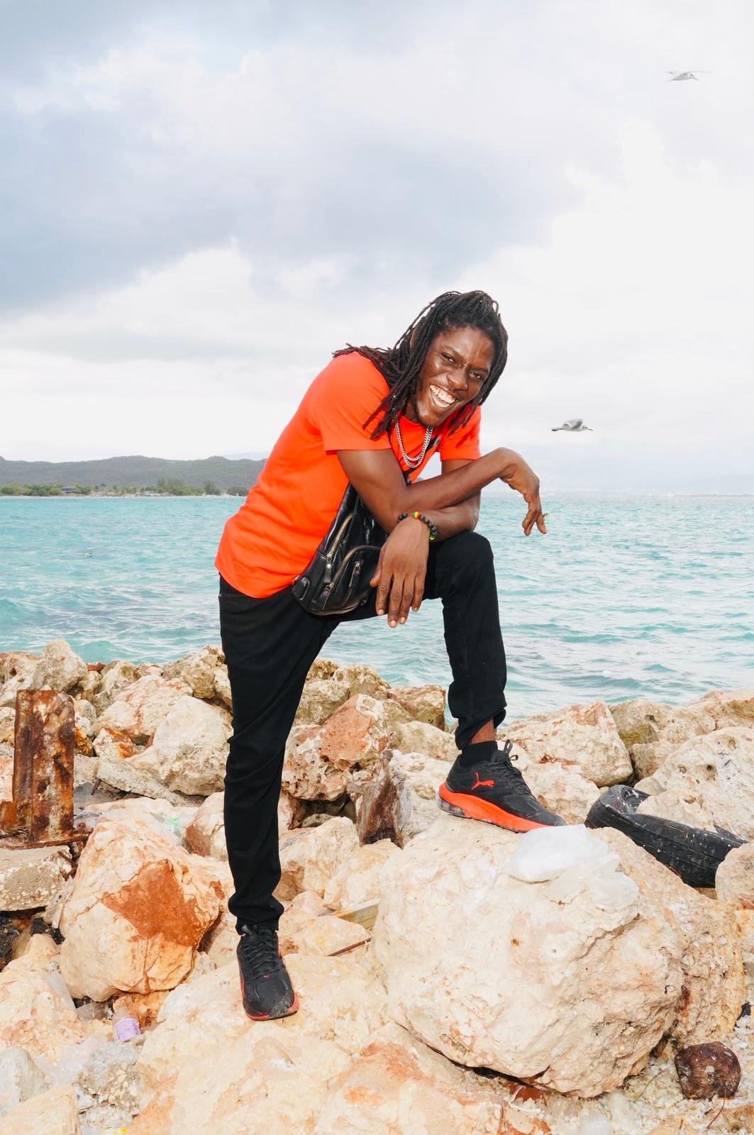 A man with dreadlocks wearing a red T-shirt, black pants, and black and red sneakers, smiling and posing on rocks by the water with an overcast sky and distant boats.