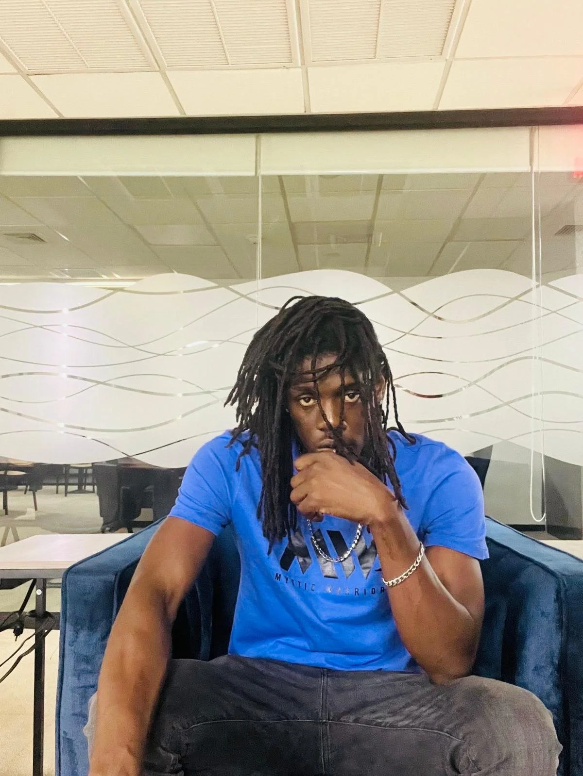 A young man with long dreadlocks wearing a bright blue t-shirt sitting on a blue velvet chair in a room with glass walls and a ceiling with white panels.