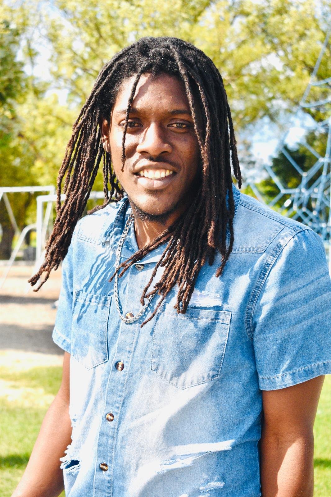 A smiling man with long dreadlocks wearing a light blue denim shirt outdoors in a park with trees and playground equipment.