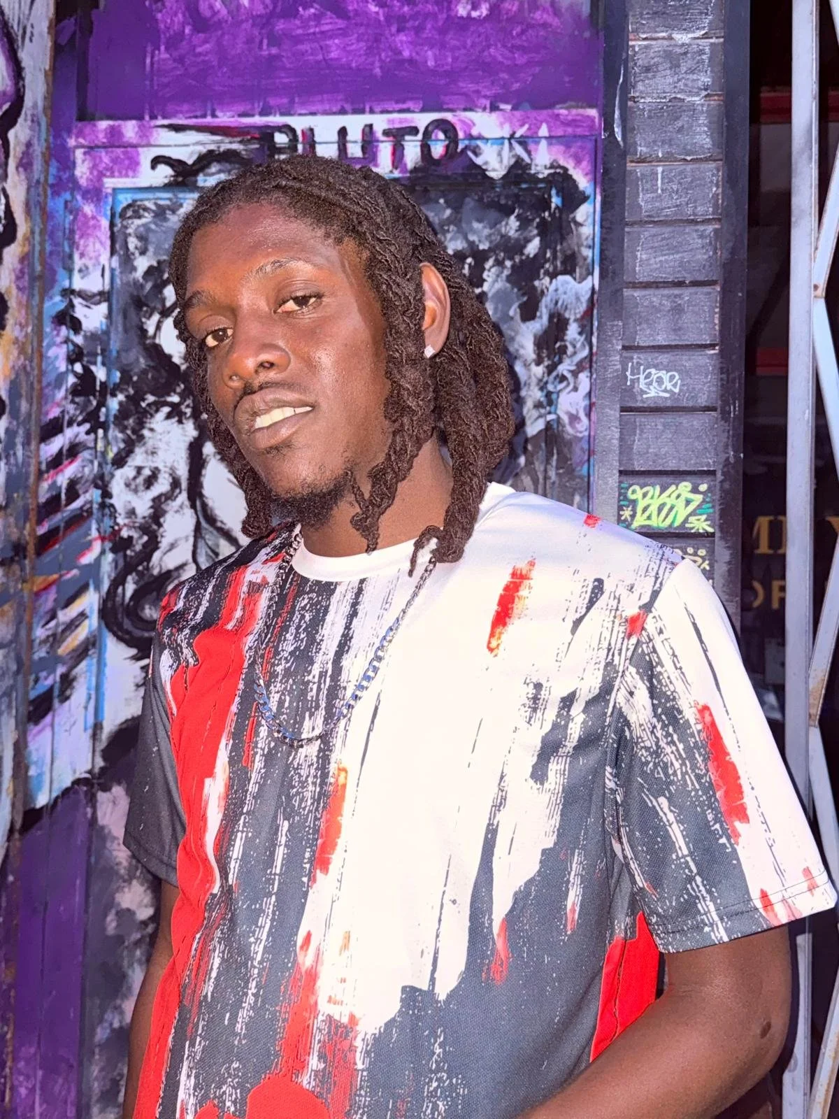 Young man with shoulder-length dreadlocks, wearing a colorful, abstract-patterned t-shirt, standing in front of a graffiti wall with purple, black, and white artwork.