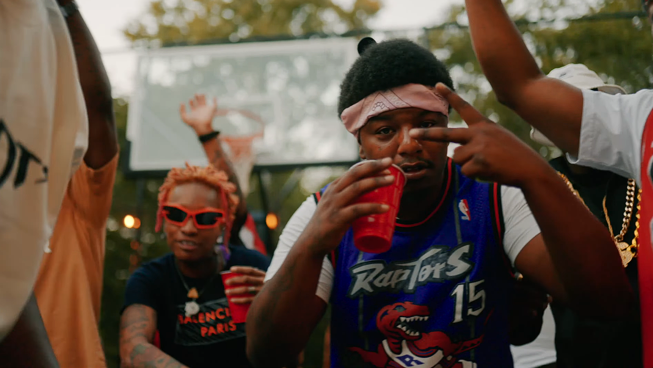 Young man wearing a Toronto Raptors basketball jersey and a headband, holding a red cup and making a peace sign, at a party with others around him, outdoors with a basketball hoop in the background.