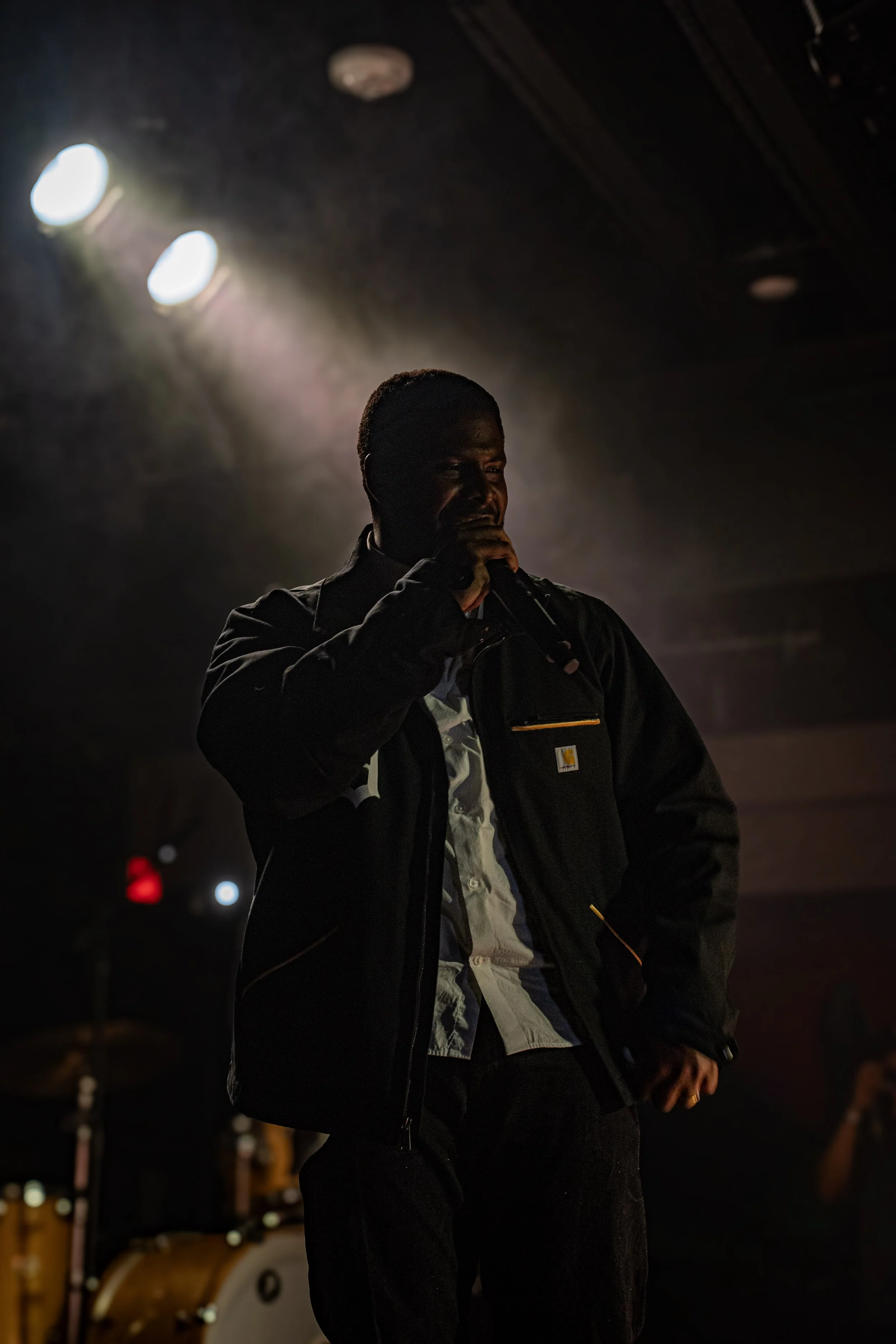 A man holding a microphone and performing on stage, with dark background and stage lights highlighting him.
