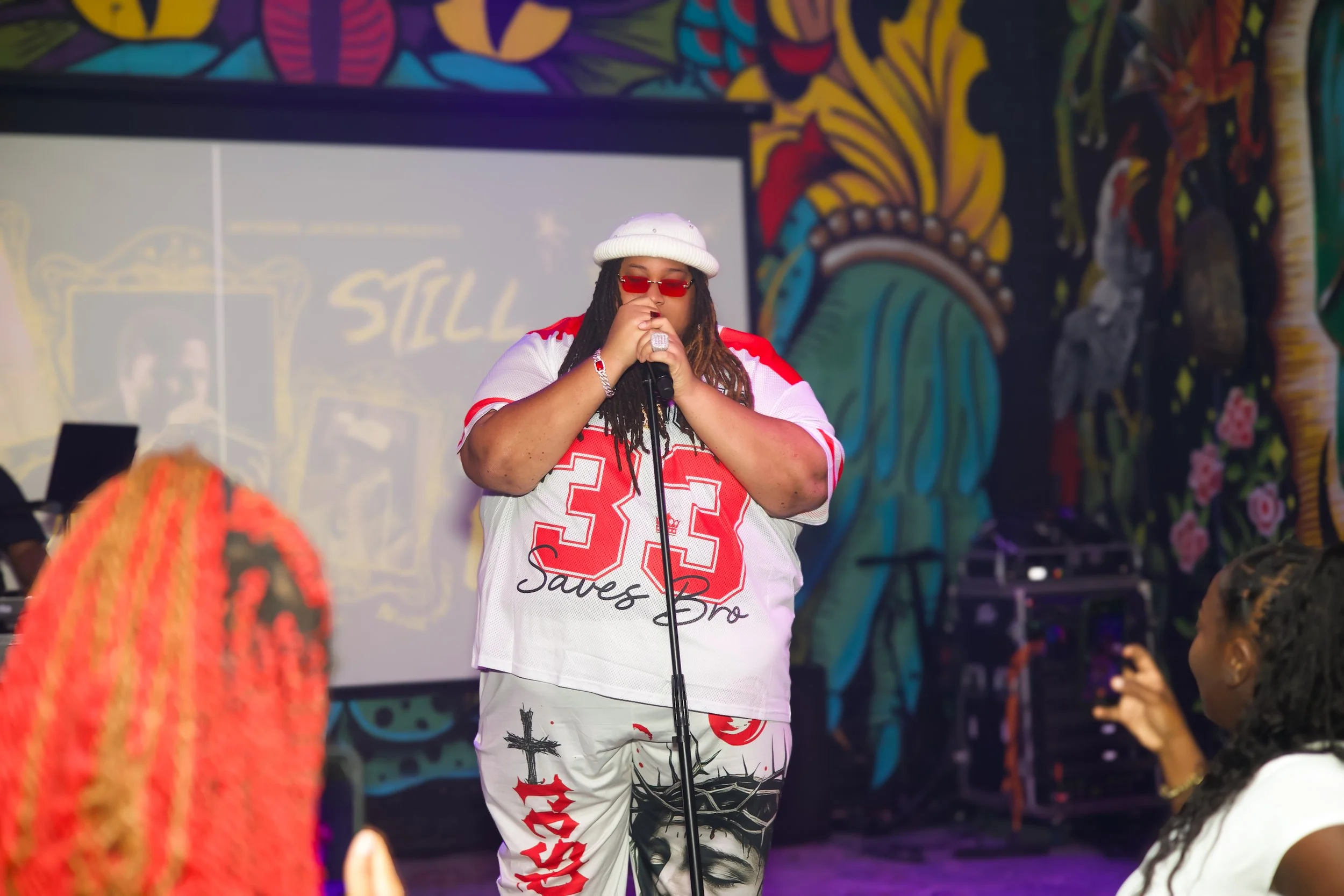 A person with long dreadlocks, wearing a white beanie, red-tinted sunglasses, and a white sports jersey with red accents and the number 33, stands on stage holding a microphone. They are in front of a colorful, graffiti-style mural, with members of t