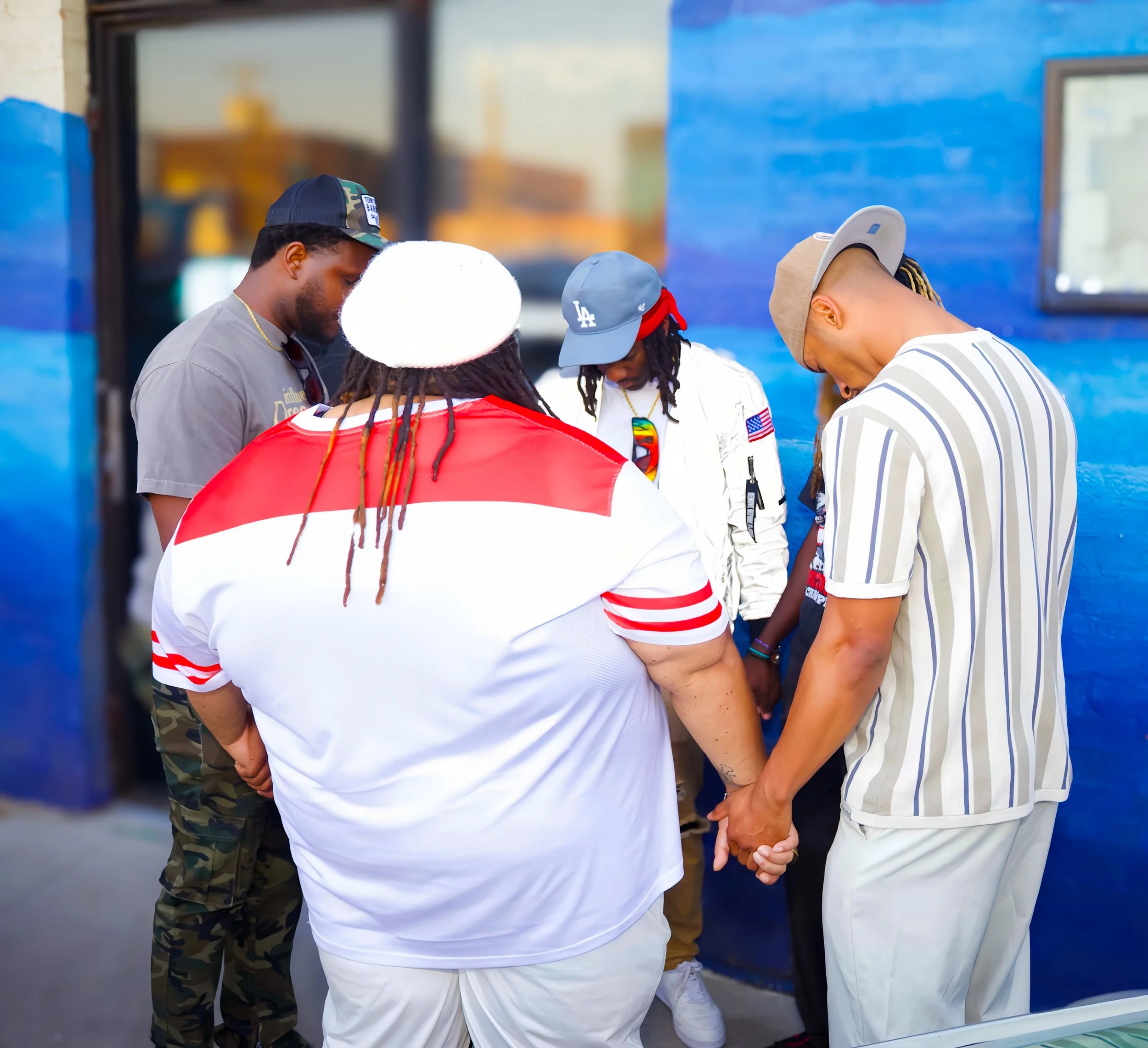 A group of people standing in a circle holding hands, with their heads bowed in prayer outdoors.