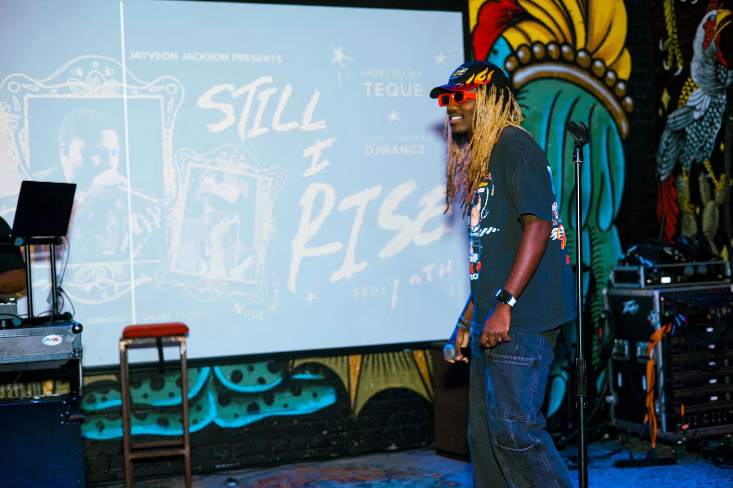 A man with long blond dreadlocks, red sunglasses, a black cap, black T-shirt with colorful graphics, and pants, standing and smiling on a stage with a microphone stand. Behind him, a vibrant wall with graffiti art, and a large screen displaying text 