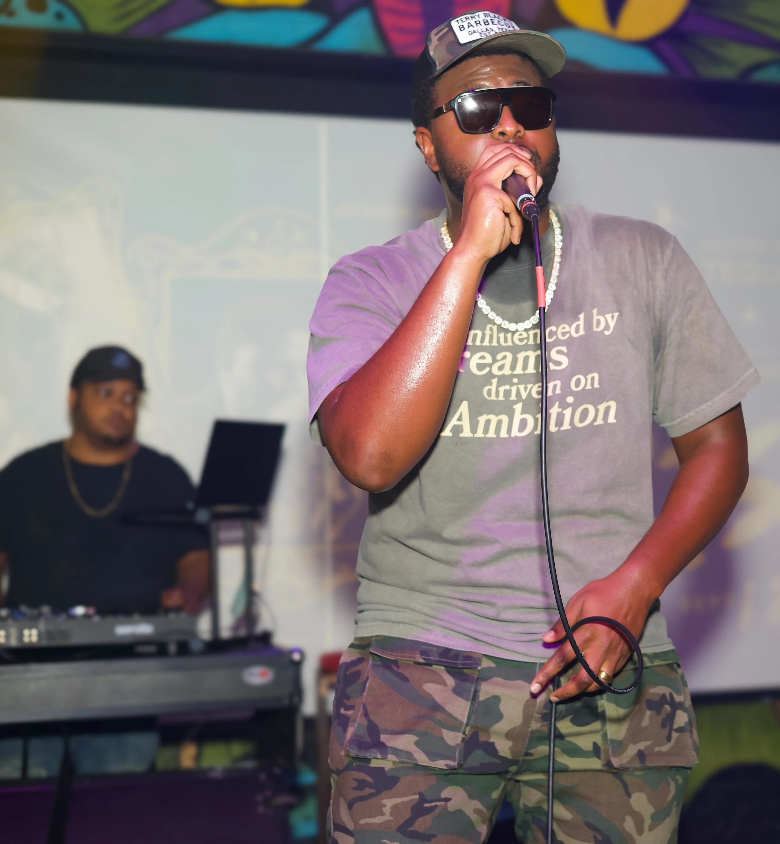 A man wearing sunglasses and a cap with a shirt that says 'Influenced by Dreams driven on Ambition' is singing into a microphone on stage. In the background, a DJ is standing behind a turntable.