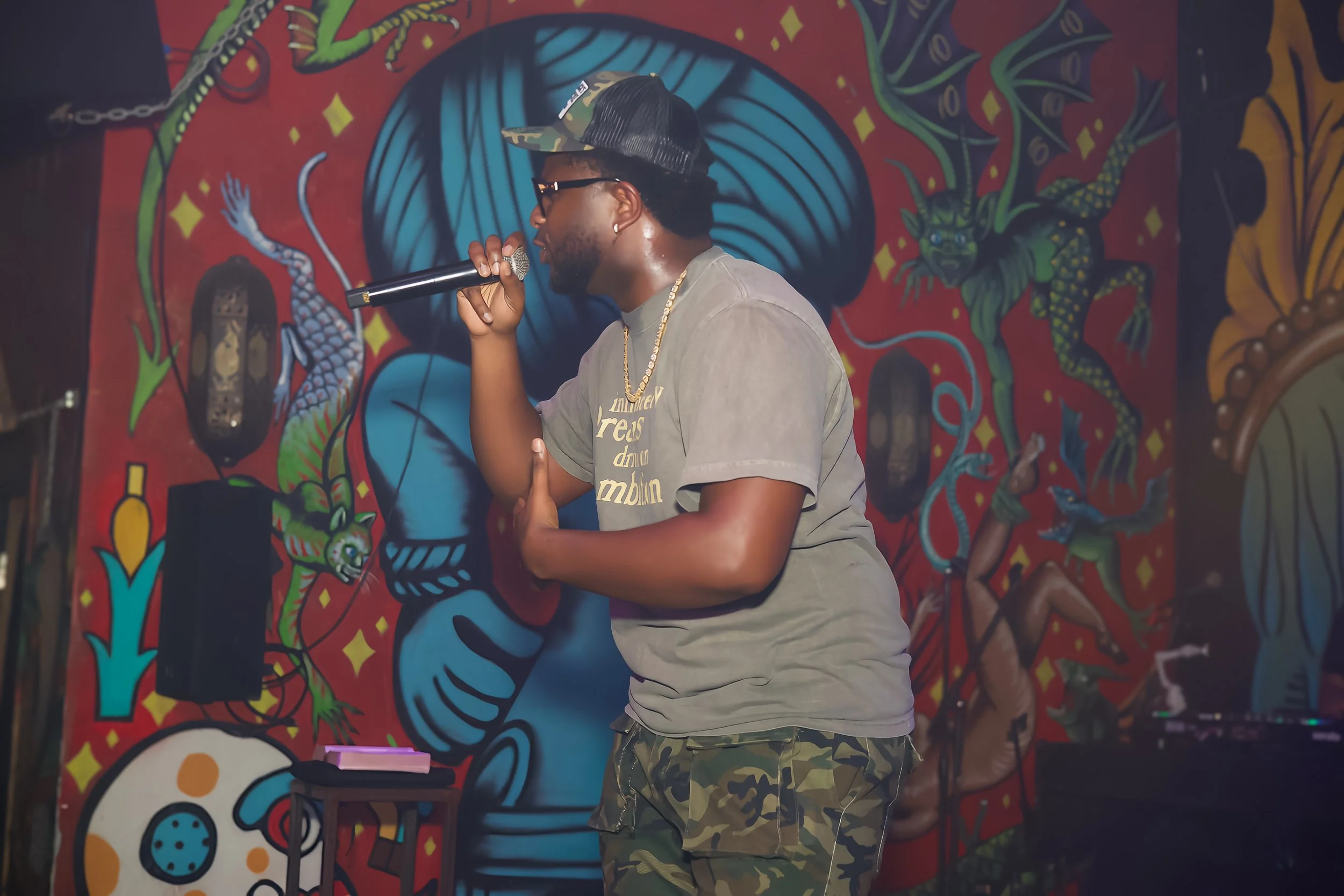 A male rapper performing with a microphone in hand in front of a colorful mural featuring dragons and other artistic designs.