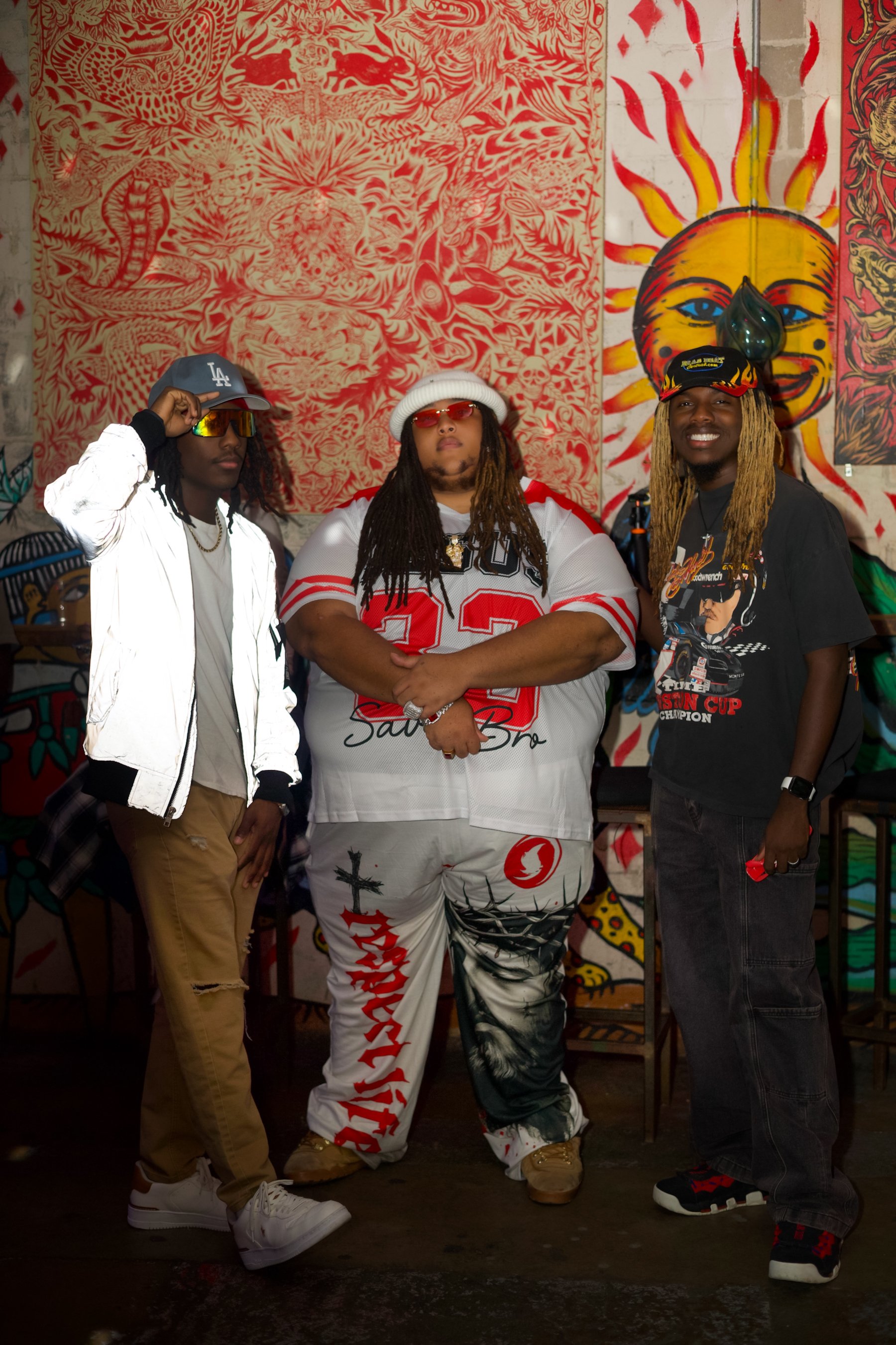 Three men standing in front of colorful graffiti art with a sun face on the wall.