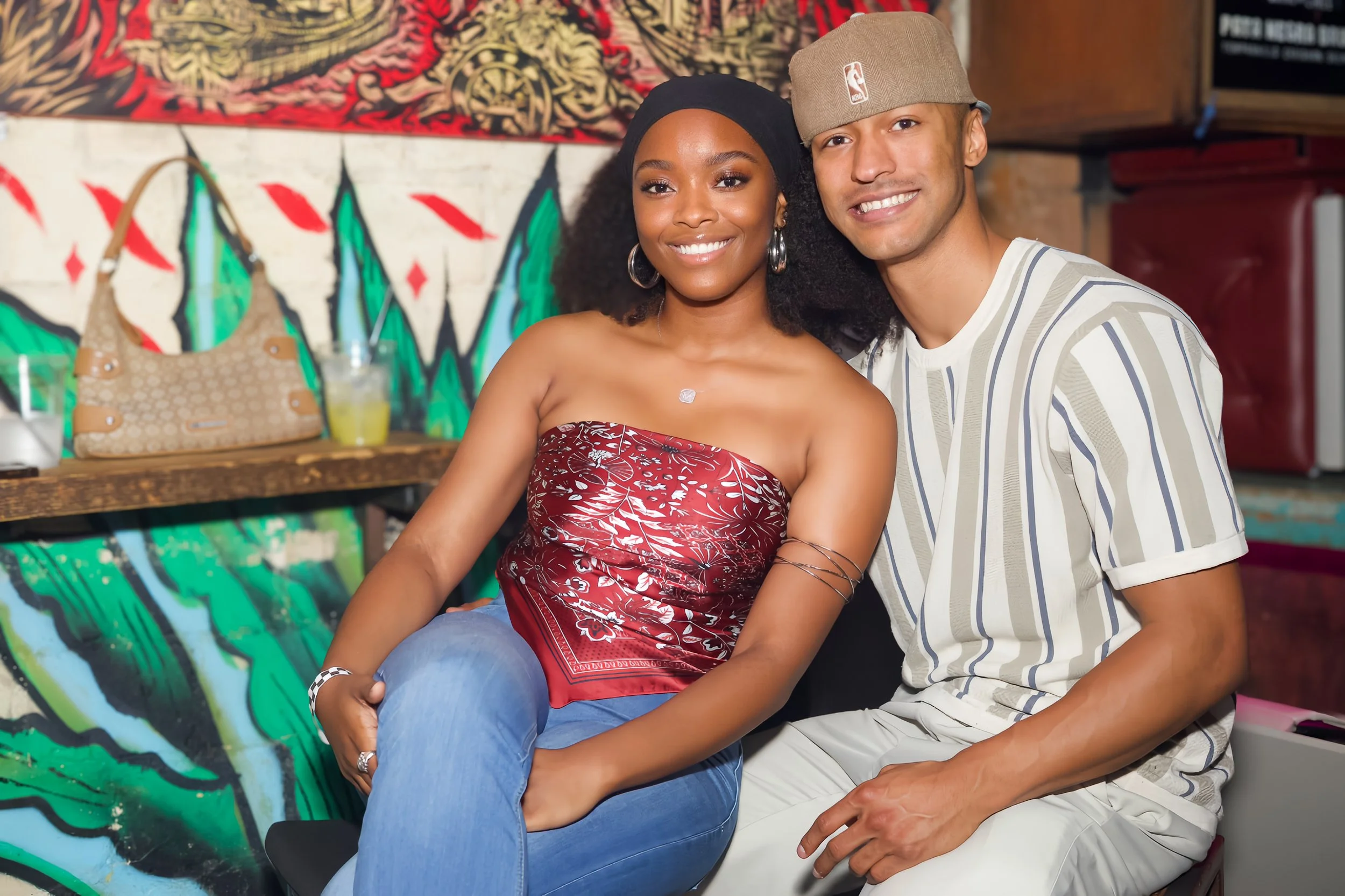 A smiling couple sitting together in front of a colorful mural, with the woman wearing a strapless red top and the man wearing a beige cap and striped shirt.