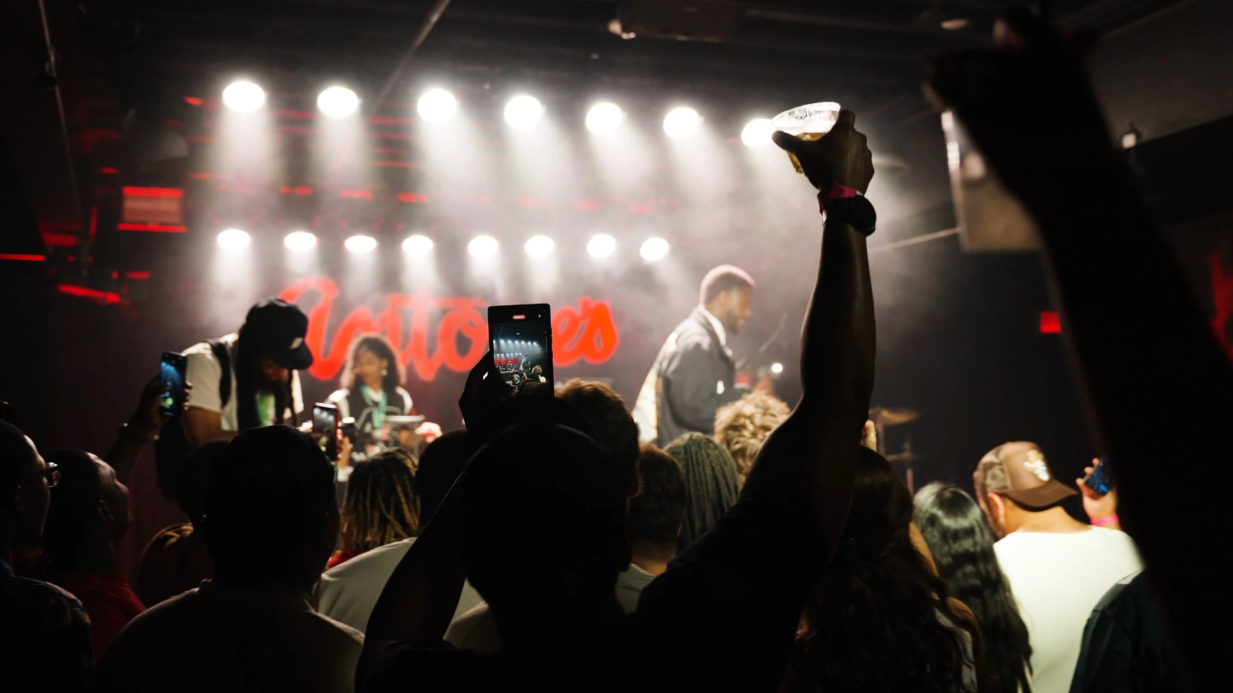 Concert crowd with audience members recording the stage on their phones, raising their hands, and enjoying live music; stage with performers and bright lights.