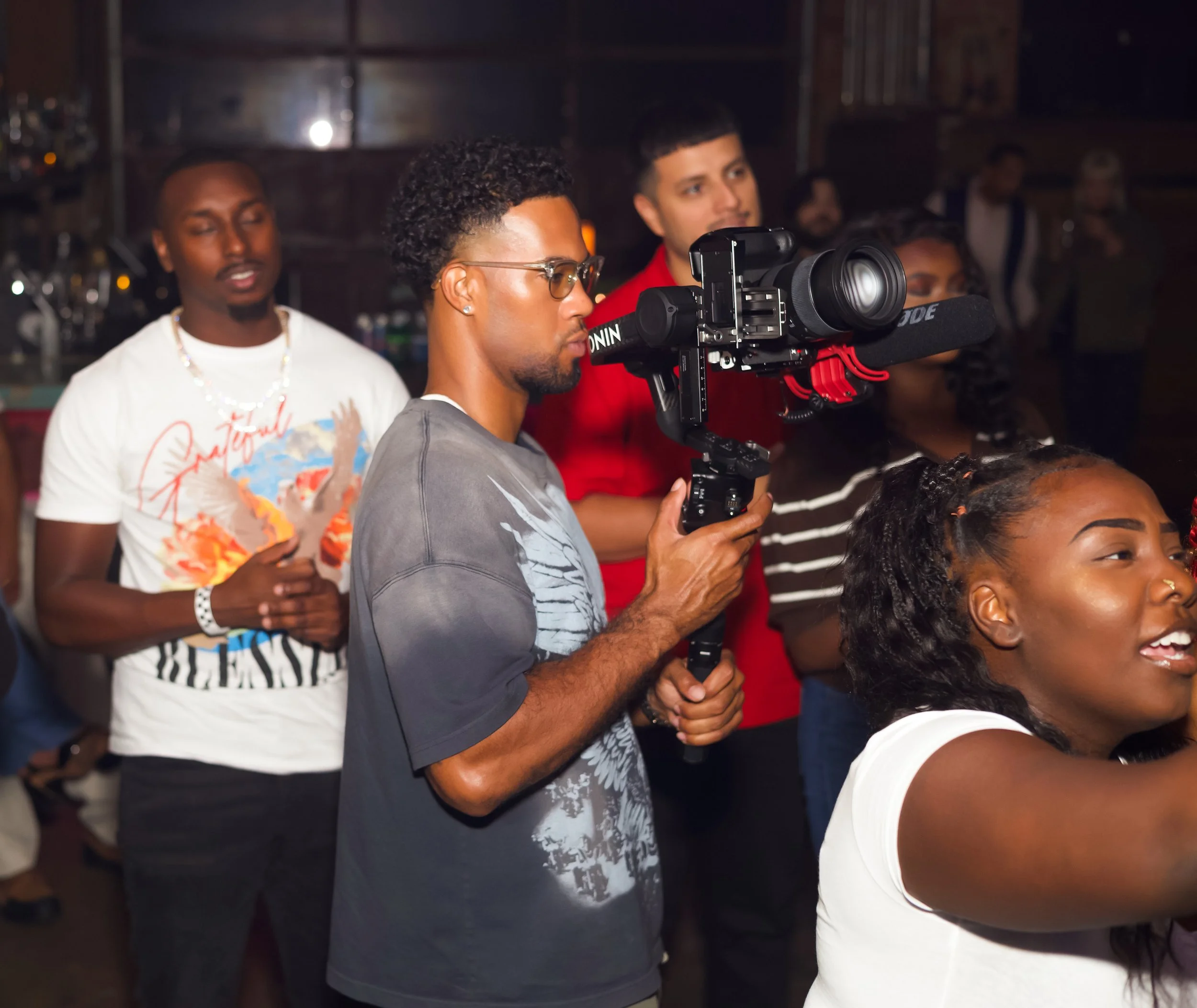 A man with glasses filming an event with a professional video camera in a dimly lit indoor setting, surrounded by other people.
