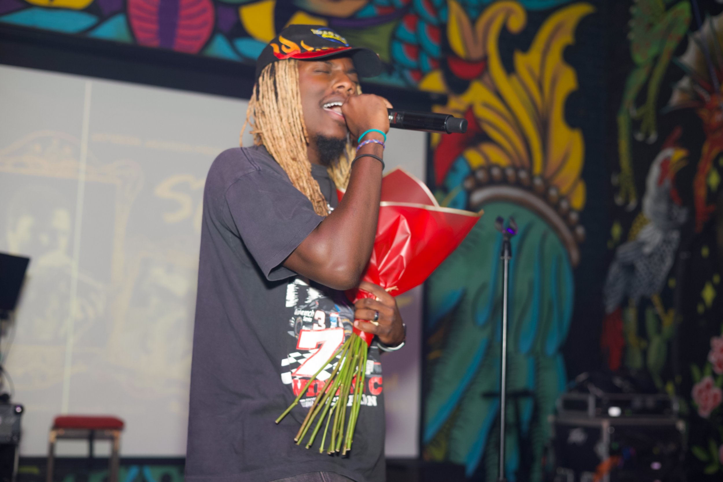 A man with long blonde dreadlocks wearing a black t-shirt and a black cap with a flame design, holding a microphone and a bouquet of red flowers, singing on stage with colorful abstract art in the background.