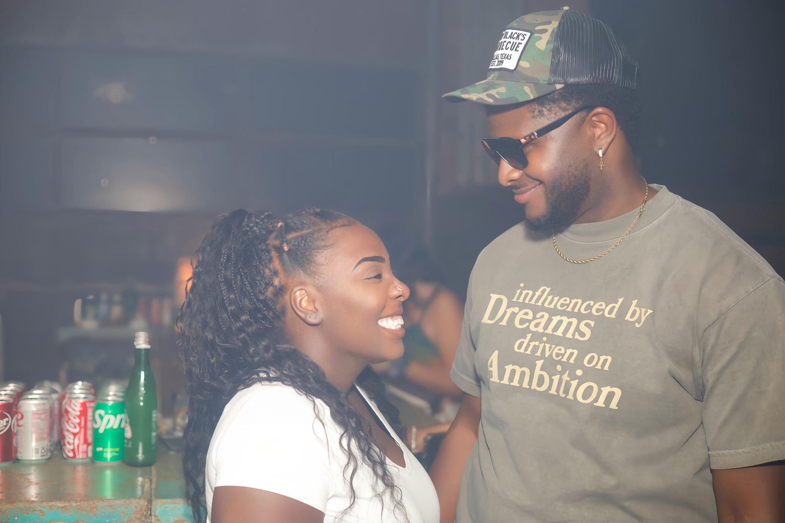 A smiling woman with braided hair and a young man wearing sunglasses, a trucker cap, and a t-shirt, standing close and looking at each other.