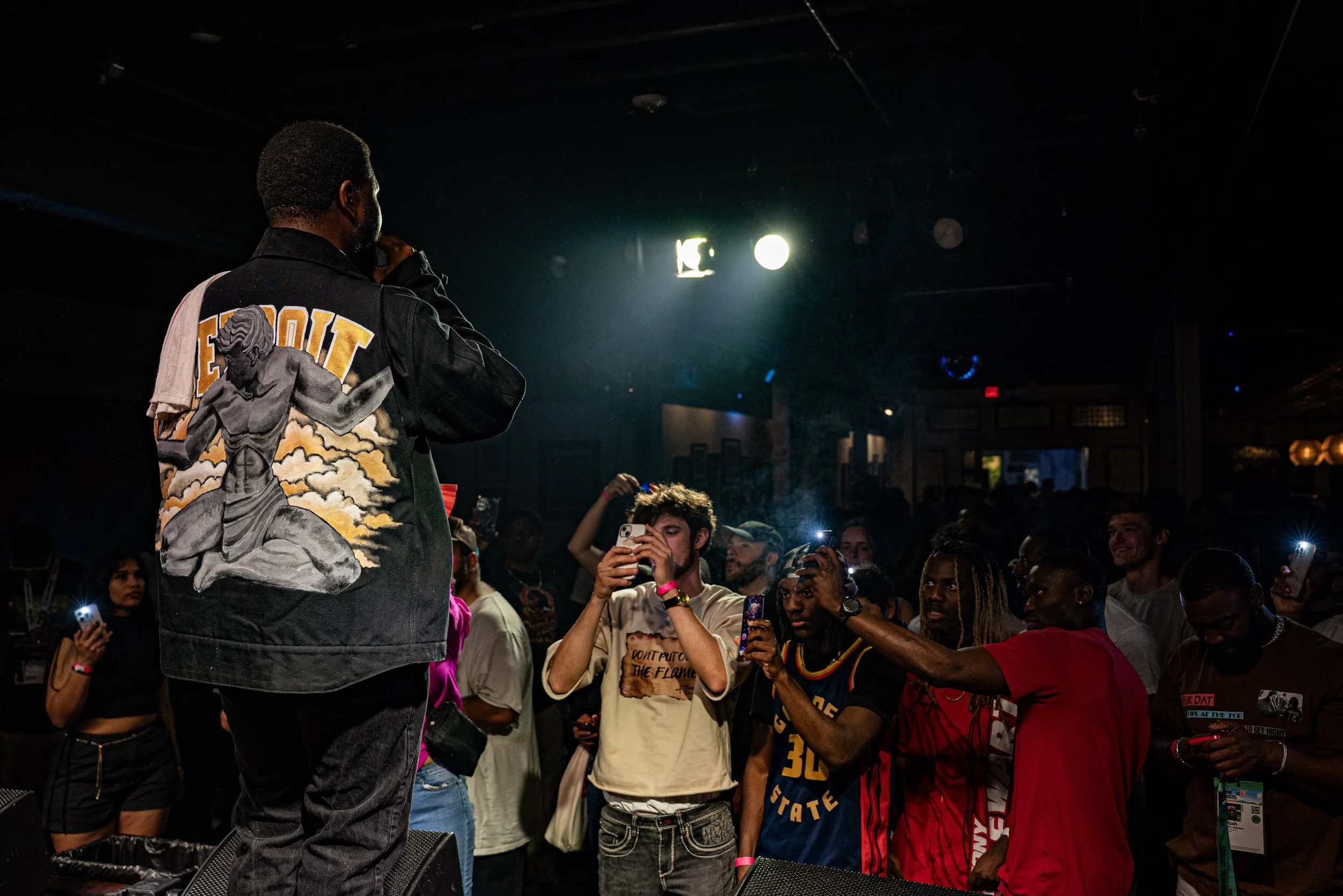 A performer on stage facing an audience at a concert or event. The performer is wearing a black jacket with a large back patch of a kneeling angel and clouds, with the word 'DETROIT' in bold yellow letters. Audience members are taking photos and videos.