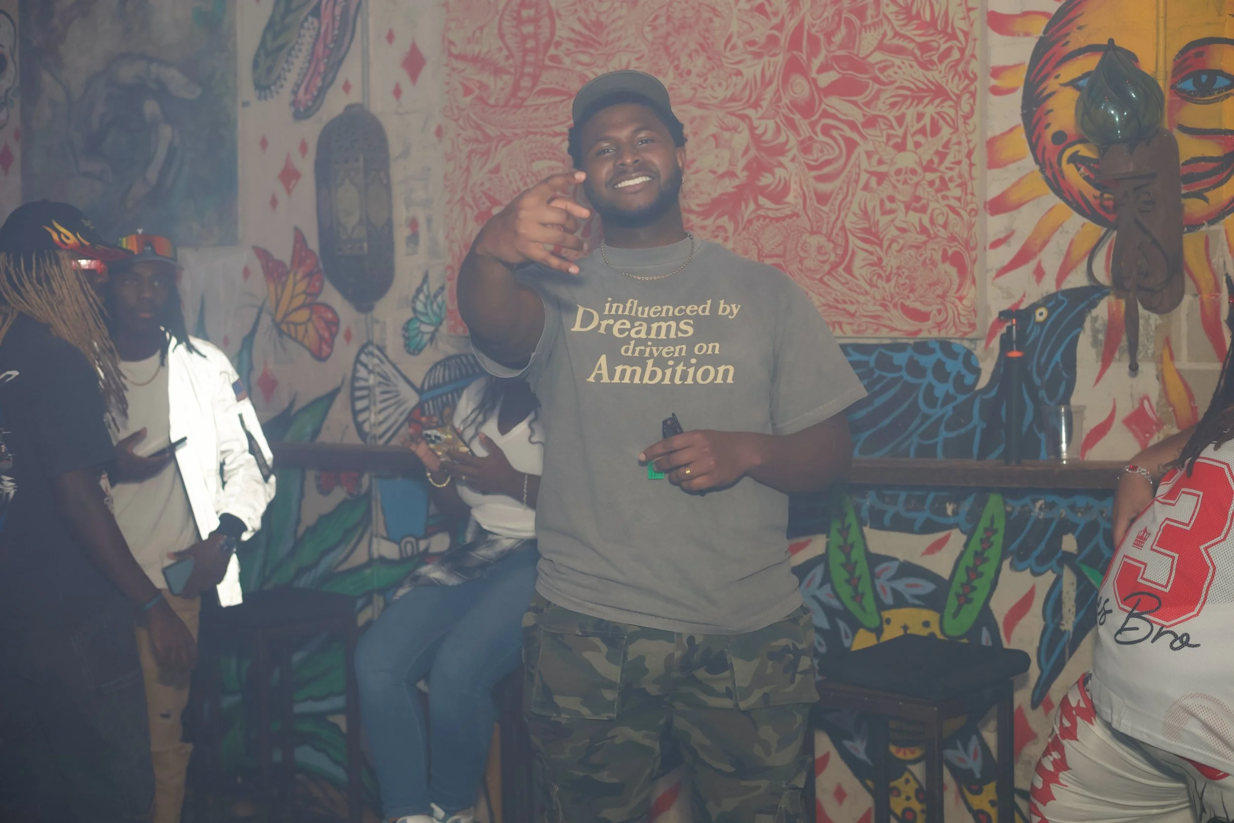 A smiling man in a gray T-shirt and camouflage shorts making a peace sign with his hand in a colorful, graffiti-style art-filled room.