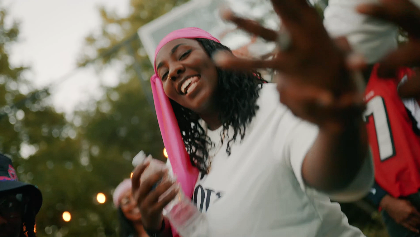 A woman wearing a pink headband and white shirt smiling and reaching toward the camera during an outdoor event, with blurred trees and string lights in the background.