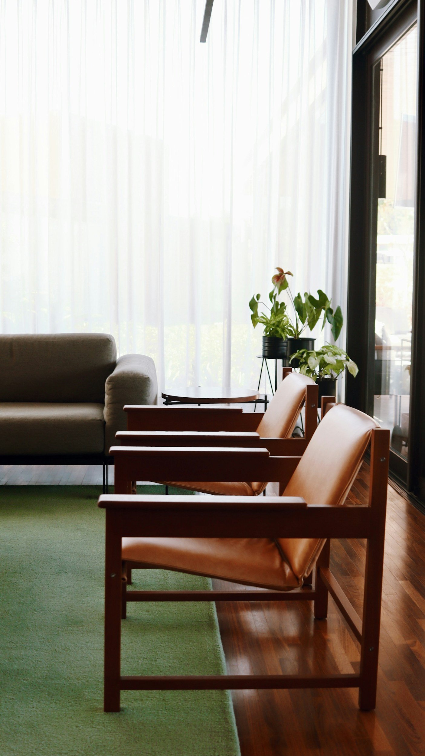 A waiting area with three wooden chairs with leather cushions, a gray sofa, a green rug, a side table, and potted plants near a large window with sheer white curtains letting in natural light.