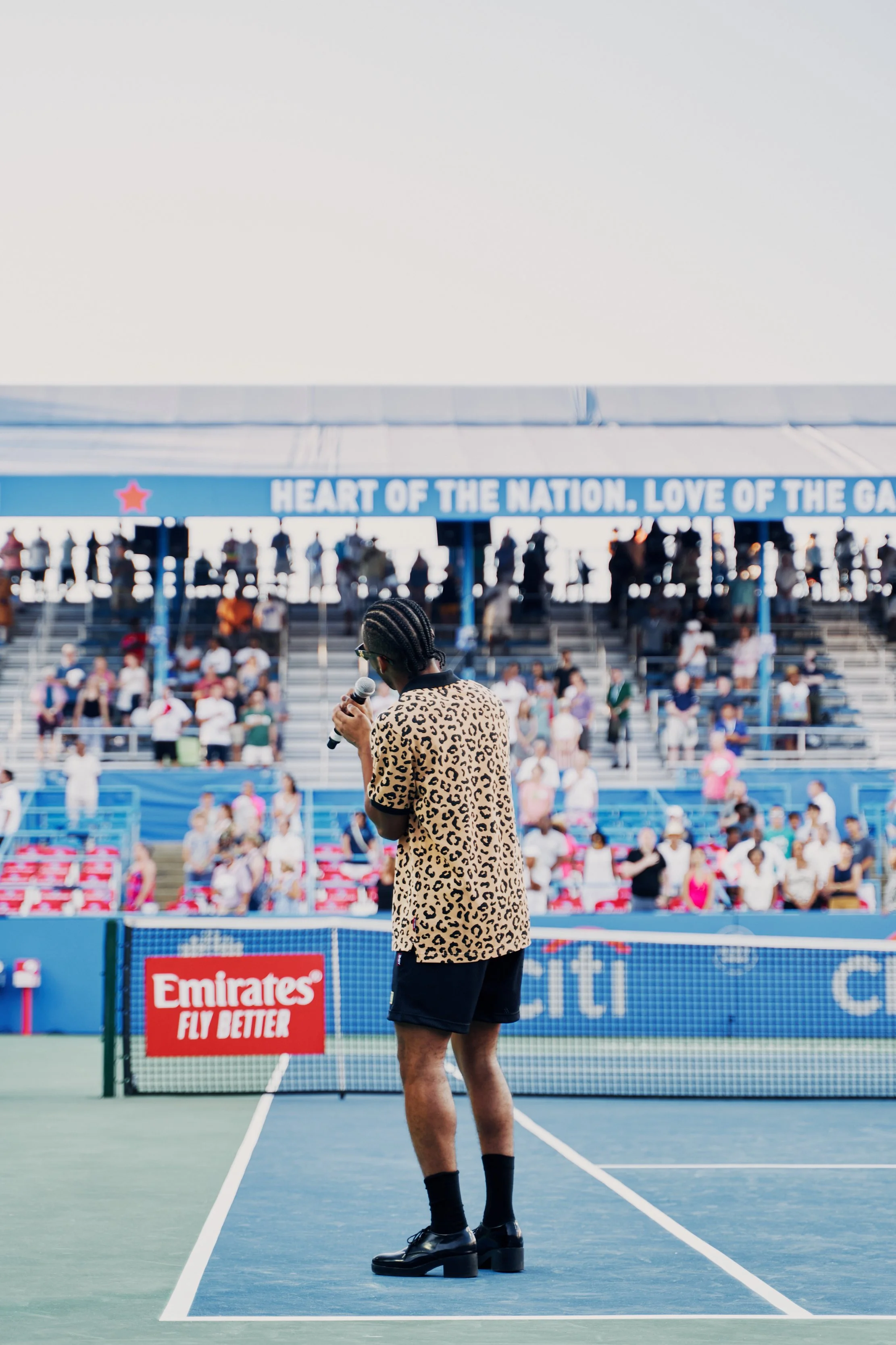 A person with braided hair wearing a leopard print shirt, black shorts, and black shoes standing on a tennis court holding a microphone, with spectators in the stands and a banner that reads 'Heart of the Nation. Love of the G' in the background.