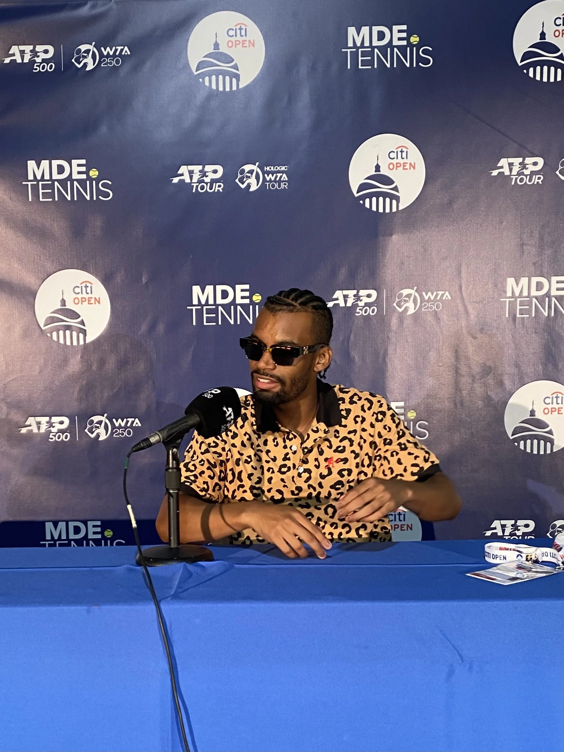 Man wearing leopard print shirt and sunglasses sitting at a blue table during a press conference at the Citi Open tennis tournament.