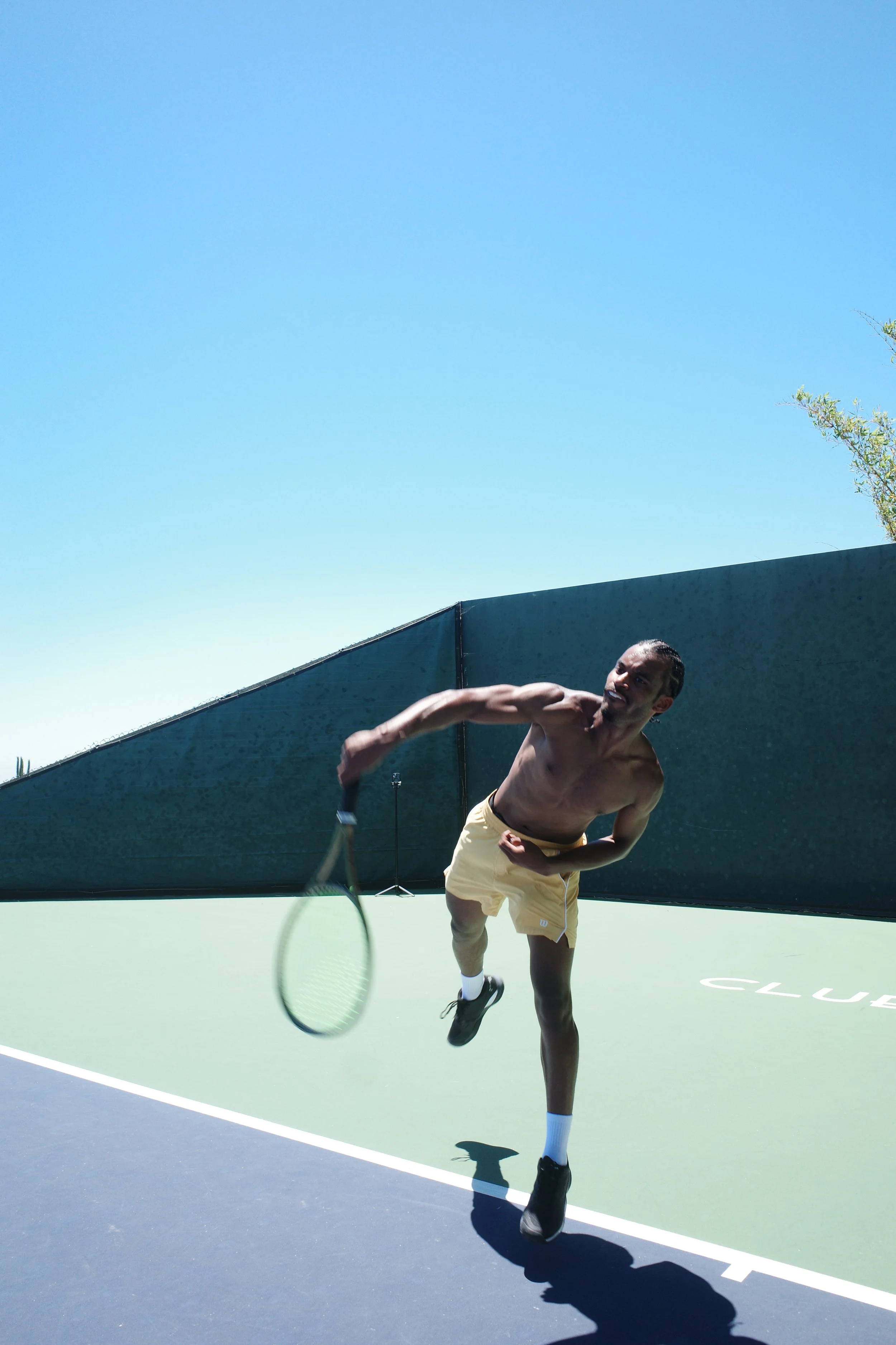 A man running shirtless on a tennis court, holding a tennis racket, with a blue sky and some trees in the background.