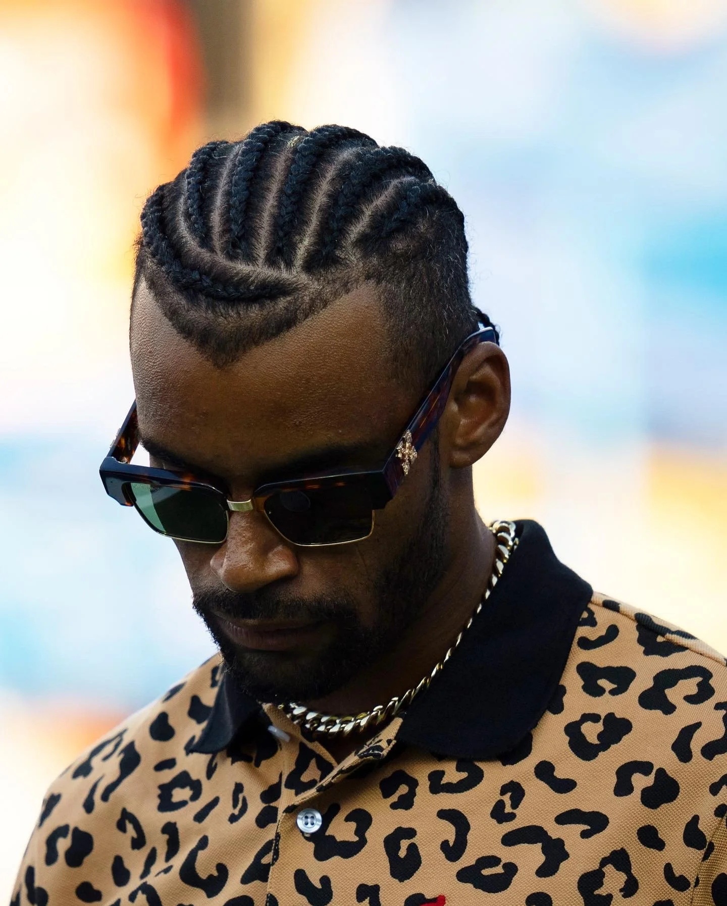 A man with cornrow hairstyle, wearing sunglasses, a gold chain, and a leopard print shirt, looking downward.