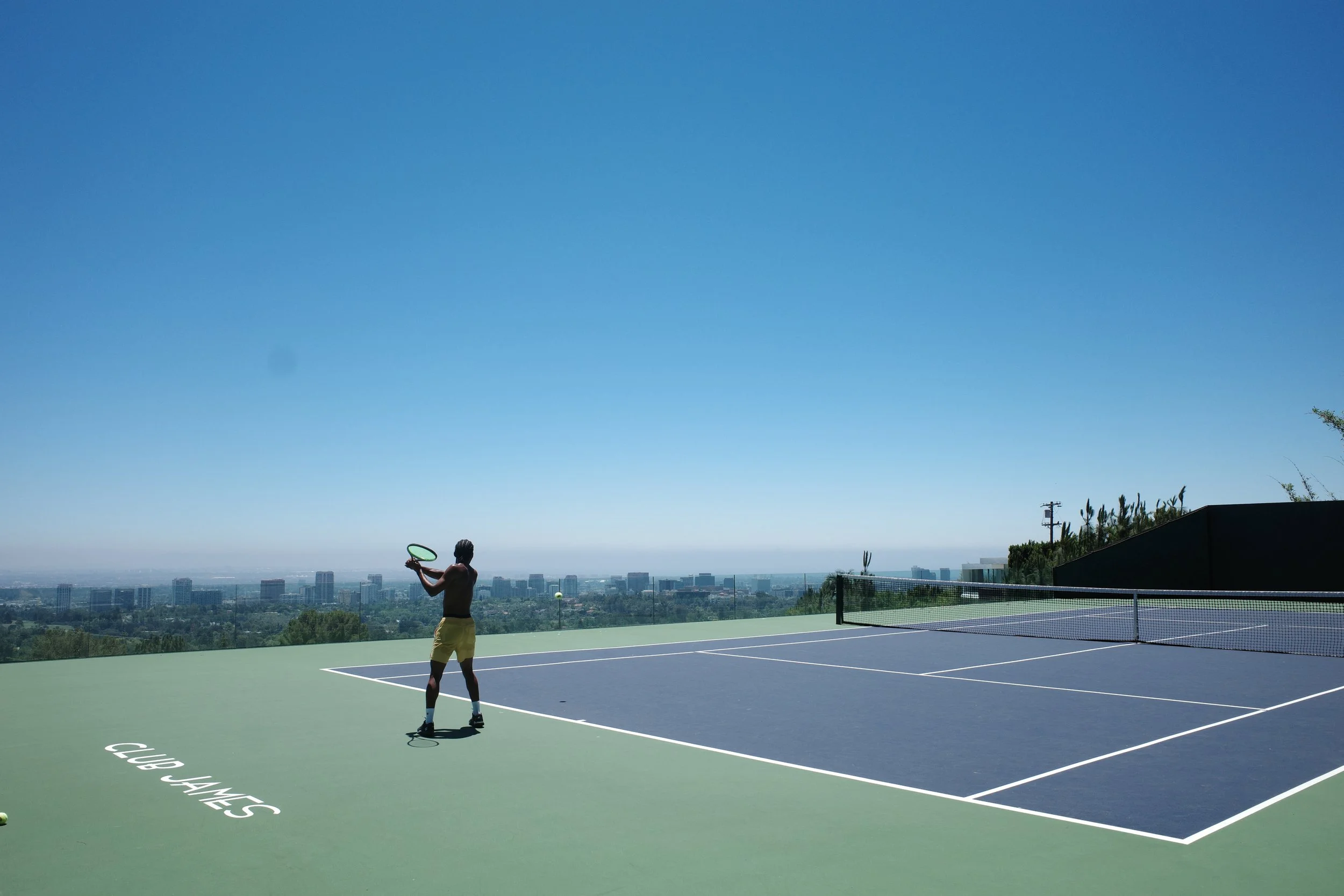 A person playing tennis on an outdoor court with a city skyline in the background under a clear blue sky.