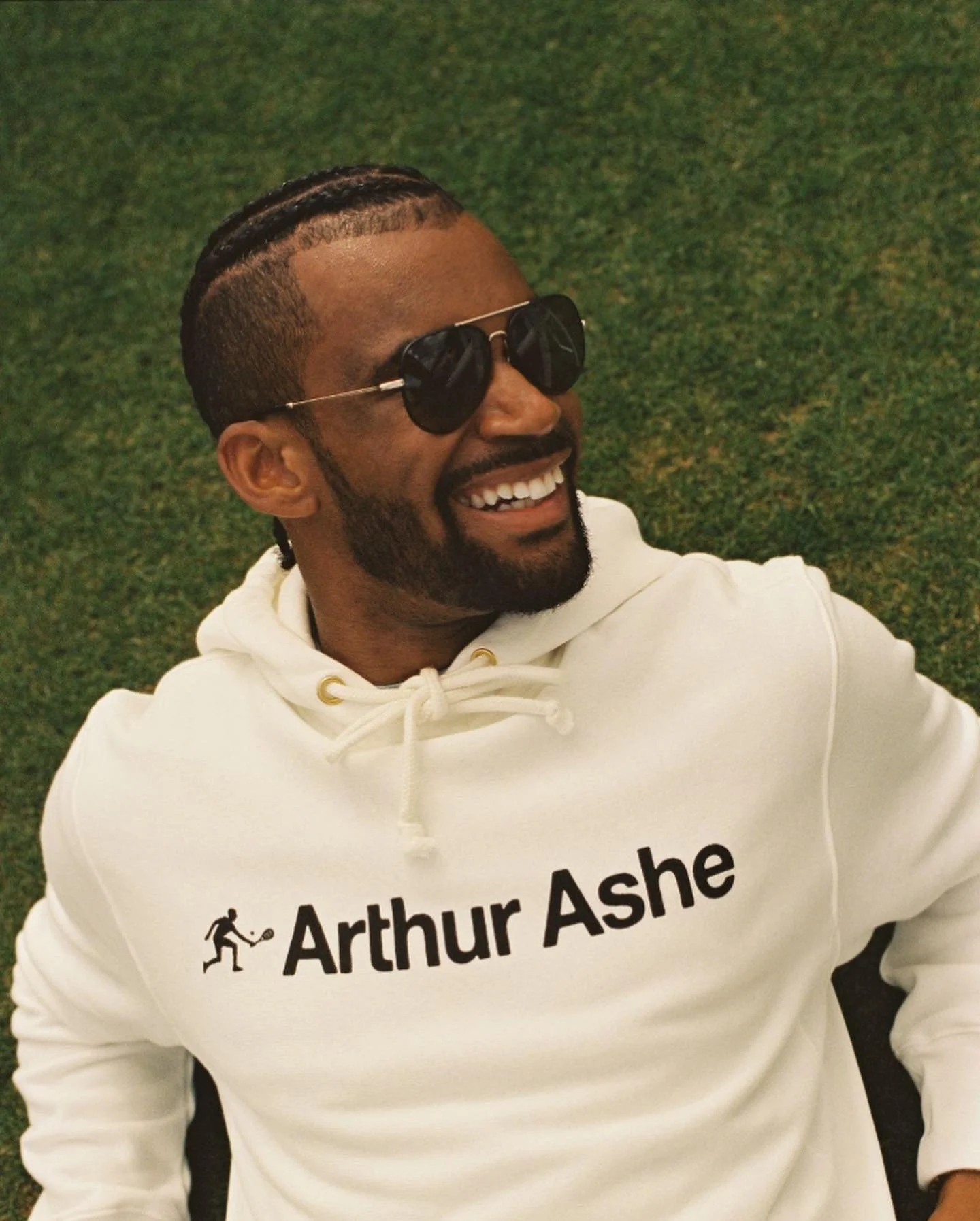 A man with braided hair, wearing sunglasses and a white hoodie with the words 'Arthur Ashe' and a tennis player icon, sitting on green grass and smiling.
