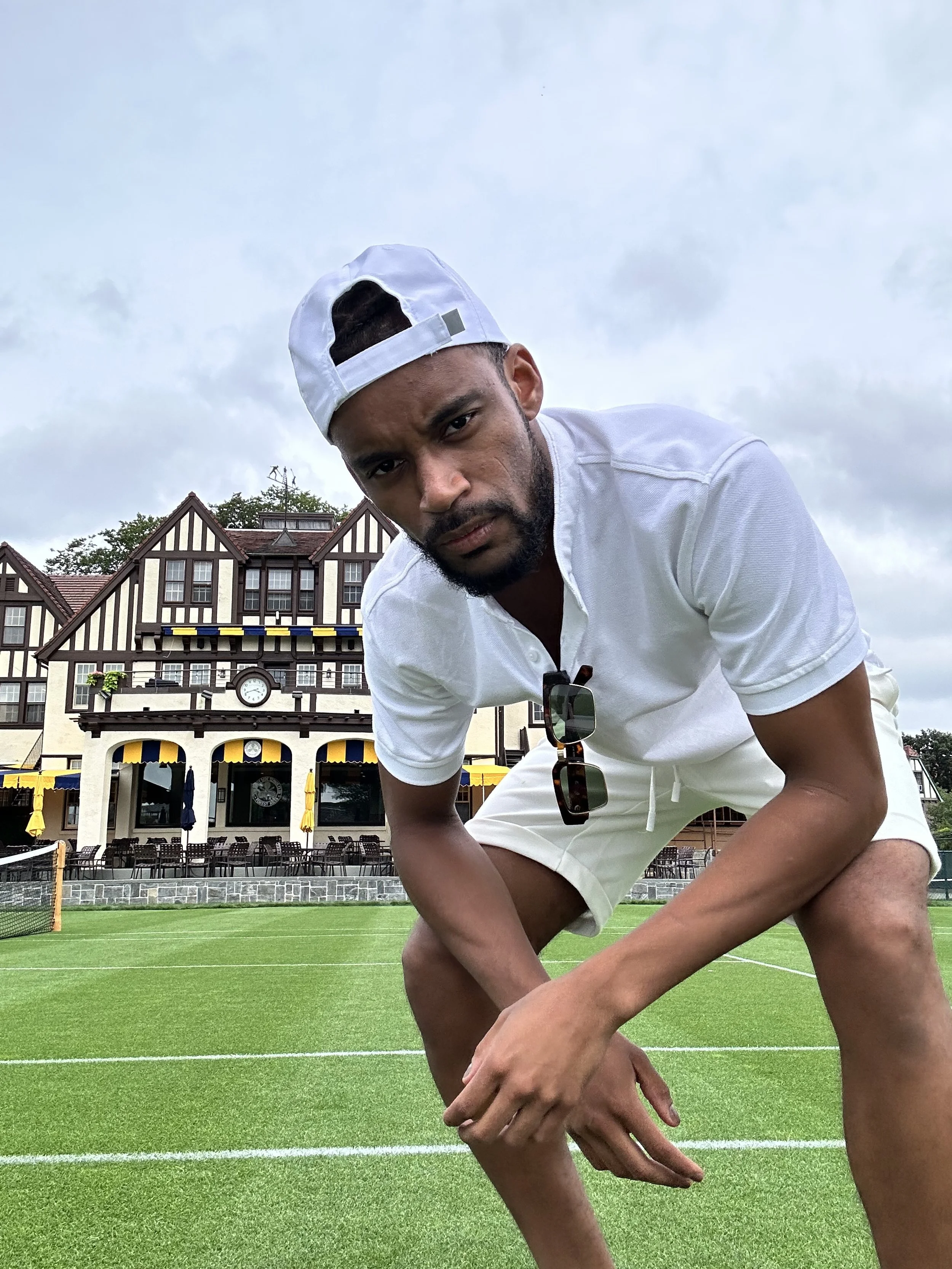 A man in a white shirt, shorts, and a backwards cap crouches on a tennis court, with a large Tudor-style building in the background.
