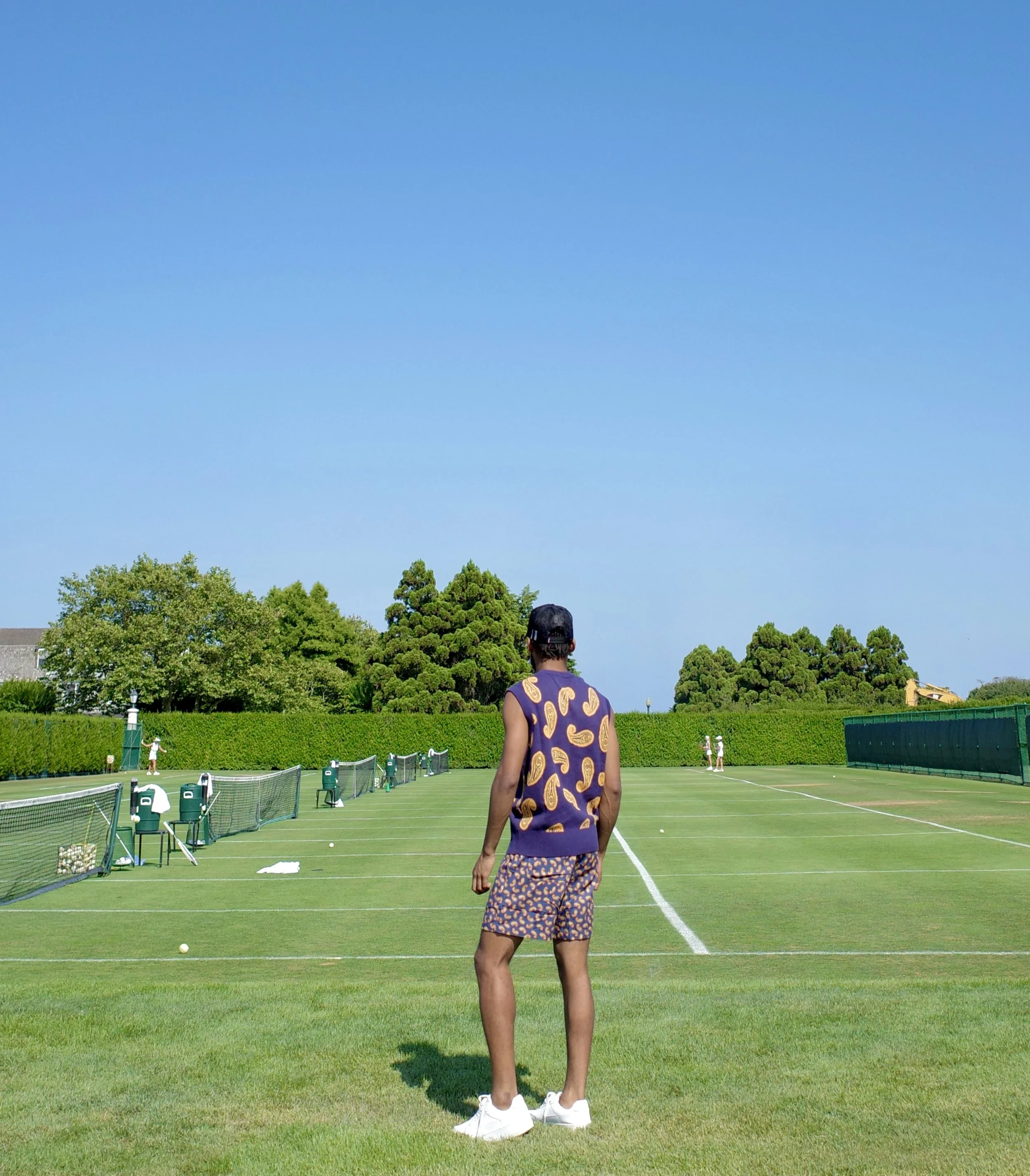 A person standing on a grass tennis court wearing a purple vest with a hot dog pattern and matching shorts, facing away from the camera under a clear blue sky.