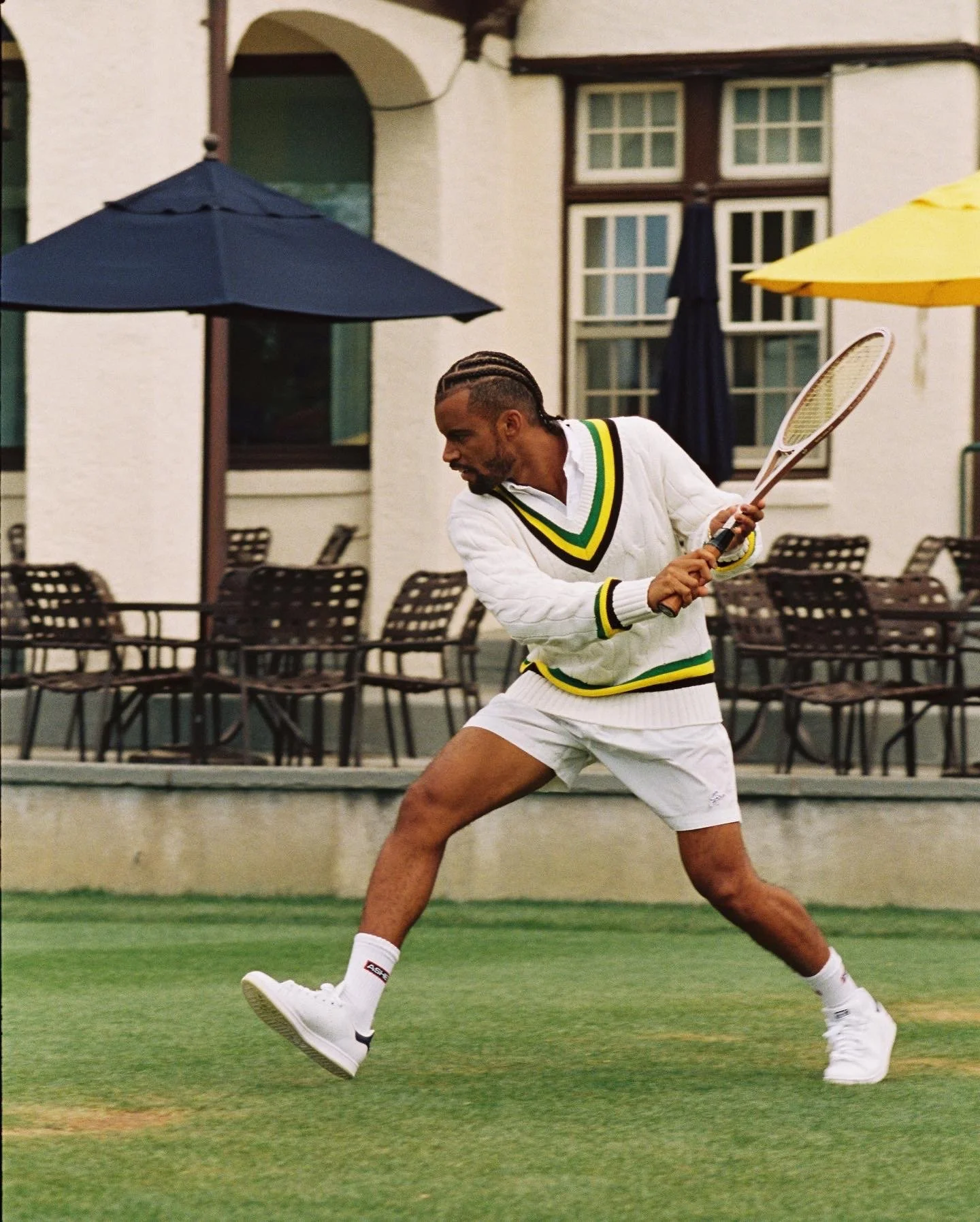A man playing tennis on a grassy court, wearing a white sweater with green, yellow, and black stripes, white shorts, white sneakers, and holding a tennis racket.