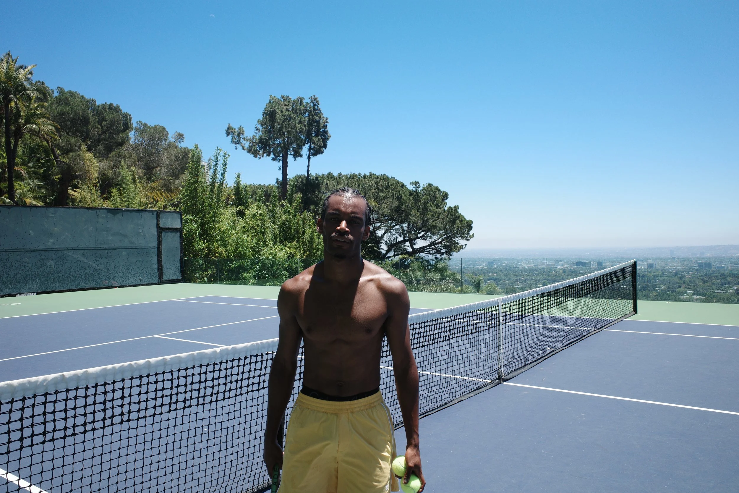 A shirtless man in yellow shorts holding tennis balls on a tennis court with a view of trees and a city in the distance under a clear blue sky.