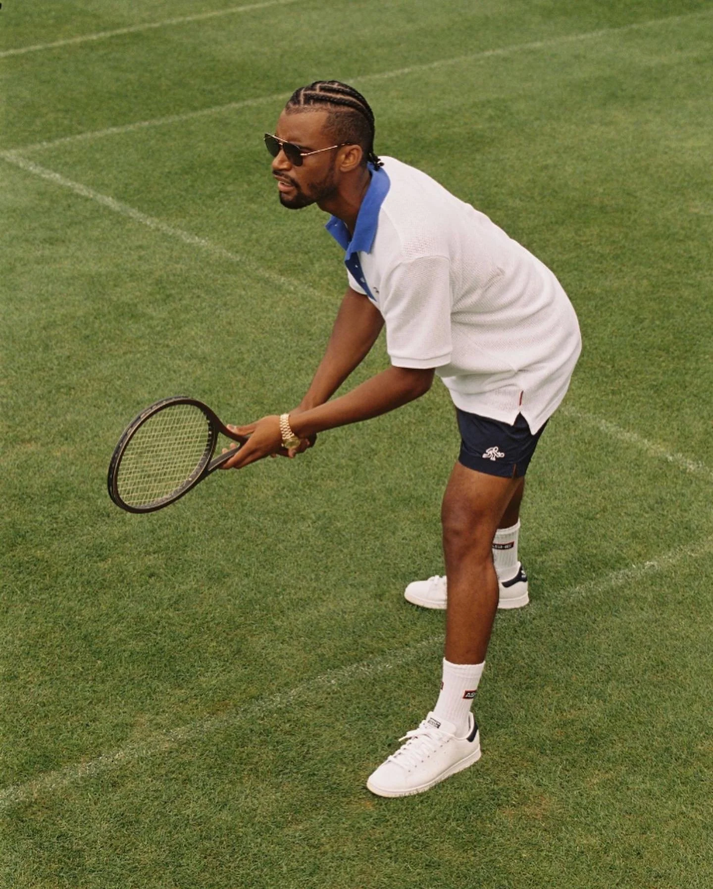 A man with braided hair, wearing sunglasses, a white and blue polo shirt, navy shorts, white sneakers, and socks, is on a tennis court holding a tennis racket in a ready position.