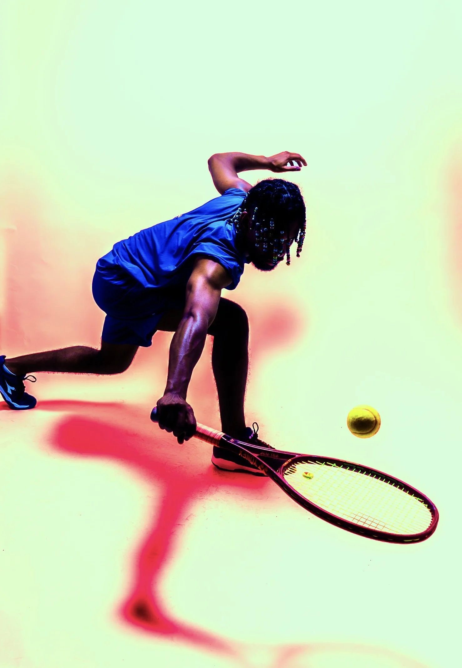 A man with dreadlocks in a blue athletic shirt and shorts is in a crouching position, holding a tennis racket and reaching toward a yellow tennis ball on a colorful background.