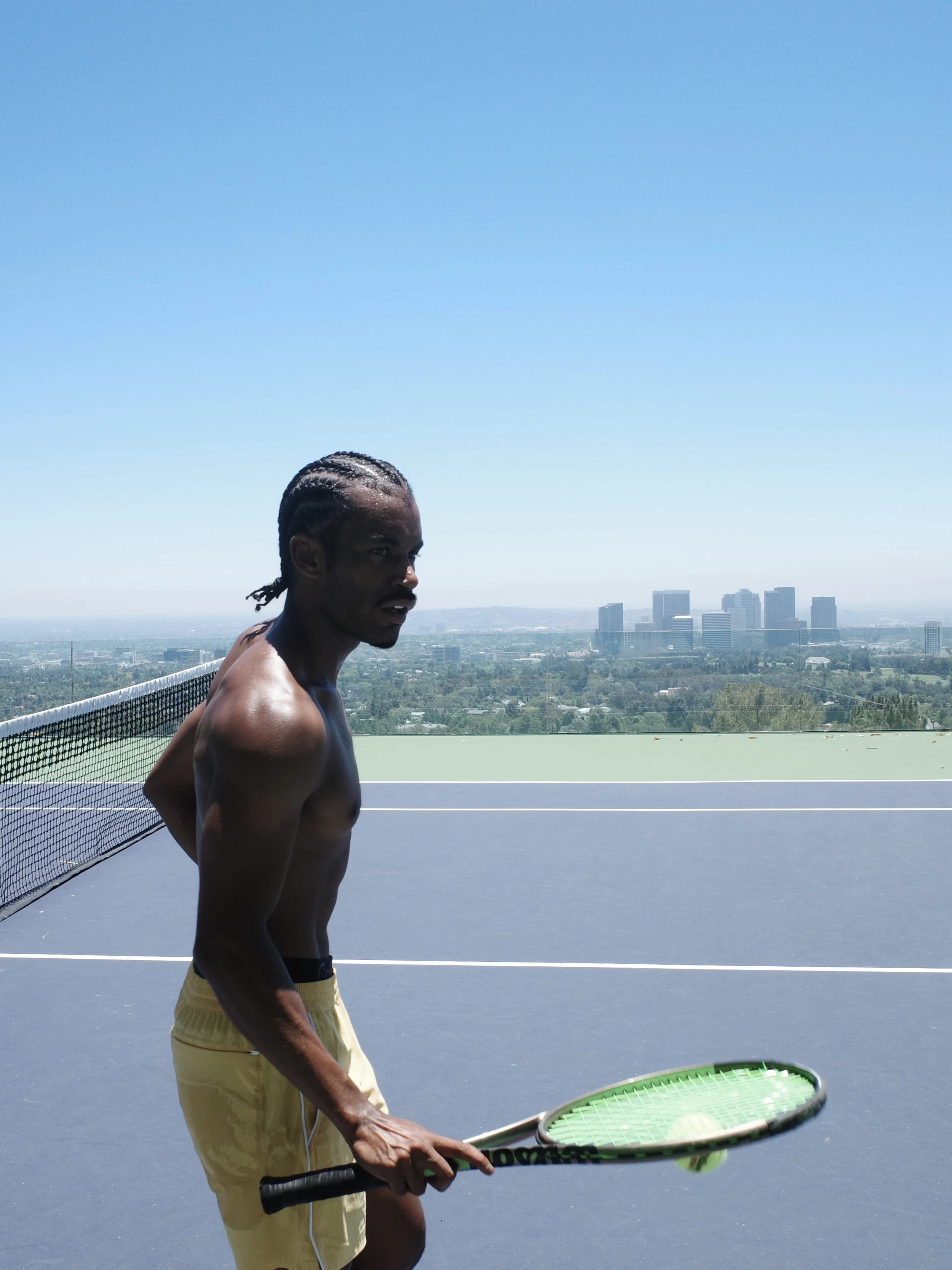 A shirtless man with cornrows holding a tennis racket on an outdoor tennis court with a city skyline in the background.