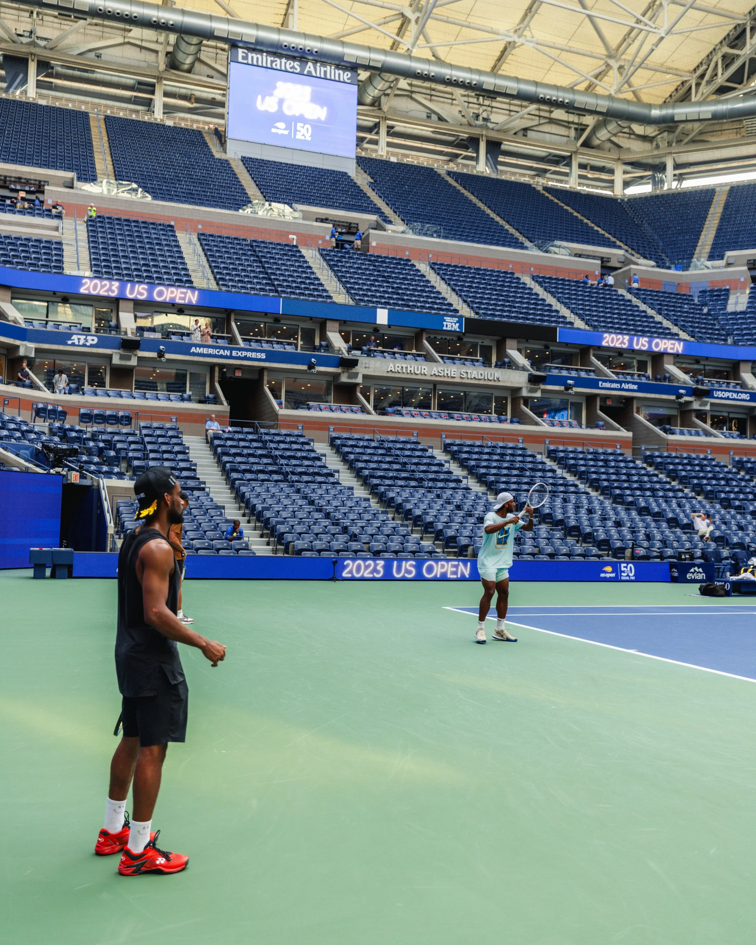 Tennis players practicing on an indoor court at the 2023 US Open, with empty spectator seats and digital banners overhead.