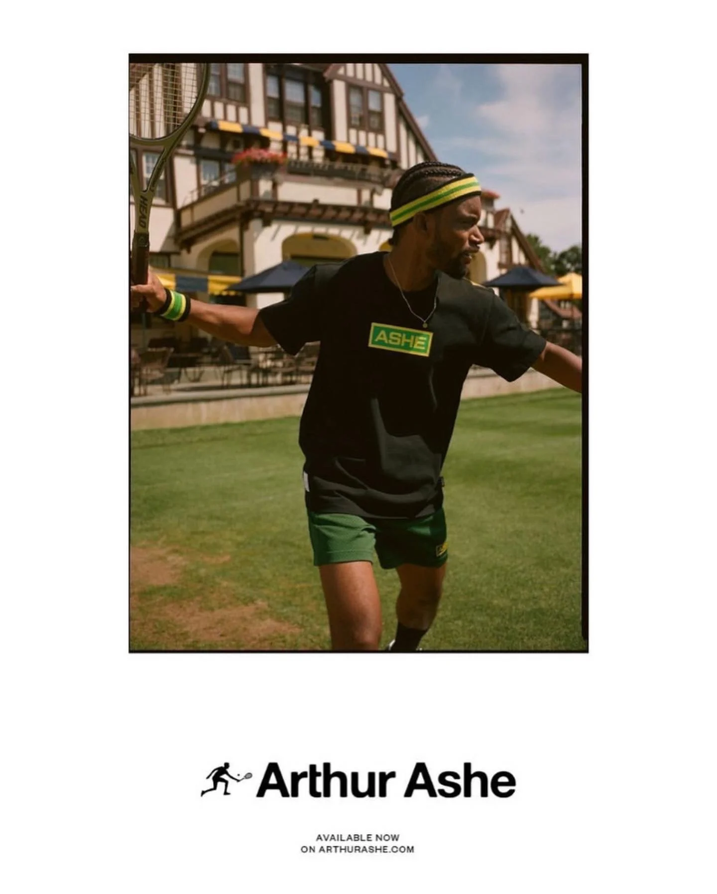 A man holding a tennis racket on a grass lawn in front of a large house, wearing a black t-shirt with 'ASHE' on it and green shorts.