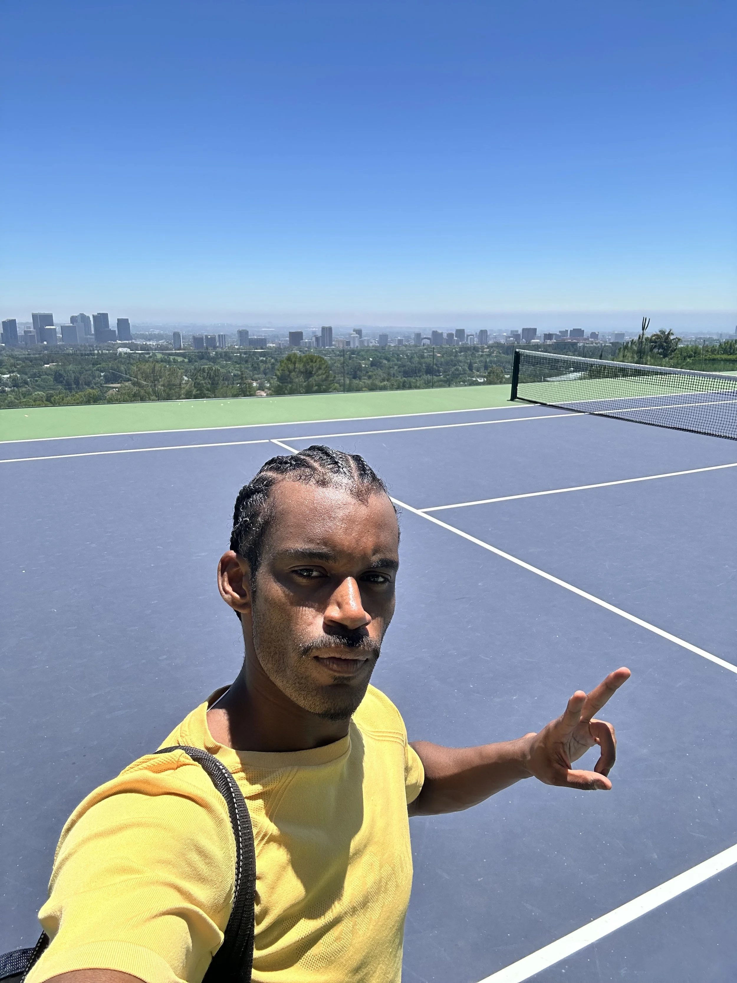 A man taking a selfie on an outdoor tennis court with a city skyline and blue sky in the background, wearing a yellow shirt and making an okay hand gesture.