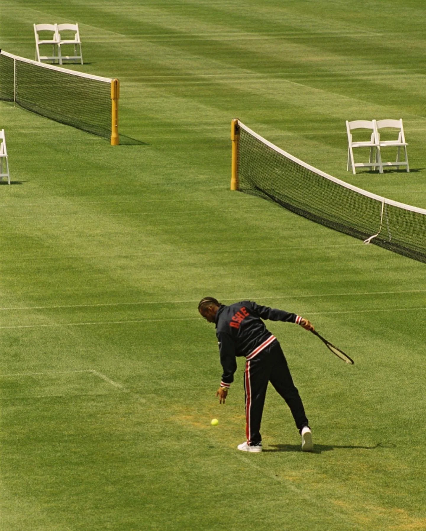 A person on a tennis court preparing to hit a tennis ball, with the court surrounded by netting and white chairs in the background.