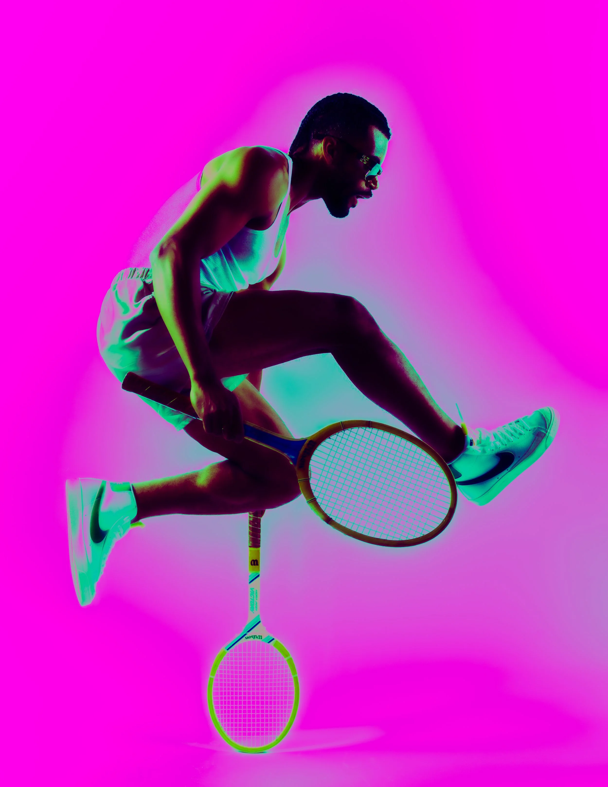 A man wearing sunglasses, shorts, and sneakers is balancing on a tennis racket, holding another tennis racket in his hand, against a bright pink background.