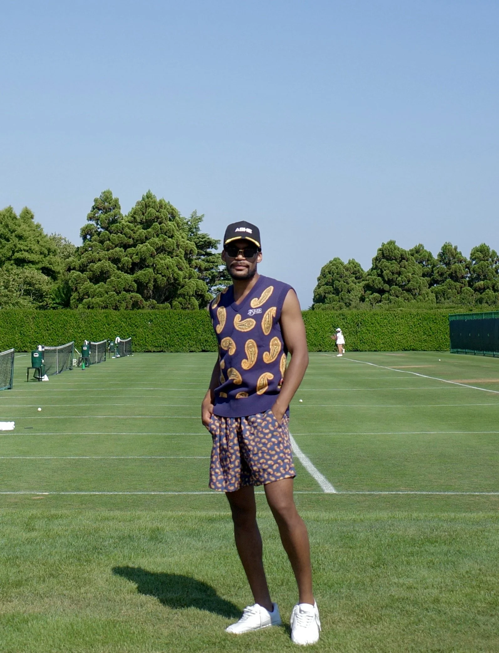 A man standing on a grass tennis court, dressed in a navy sleeveless shirt with gold paisley patterns, shorts with a matching pattern, white sneakers, sunglasses, and a cap. There are trees and tennis nets in the background.