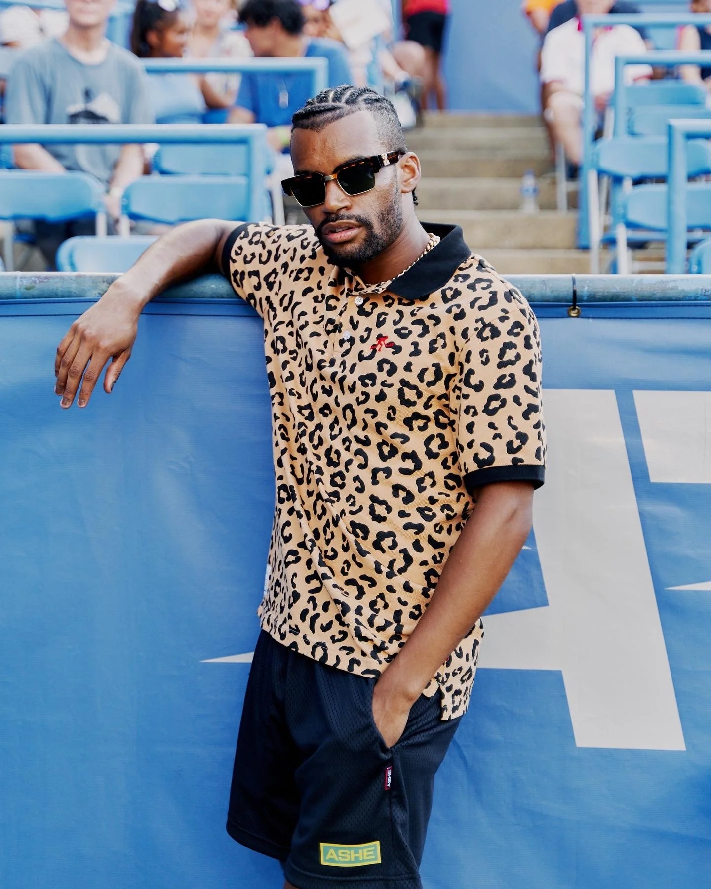 Man with braided hair wearing sunglasses, a leopard print polo shirt, and black shorts leaning against a blue barrier at an outdoor event, with people sitting on blue bleachers in the background.