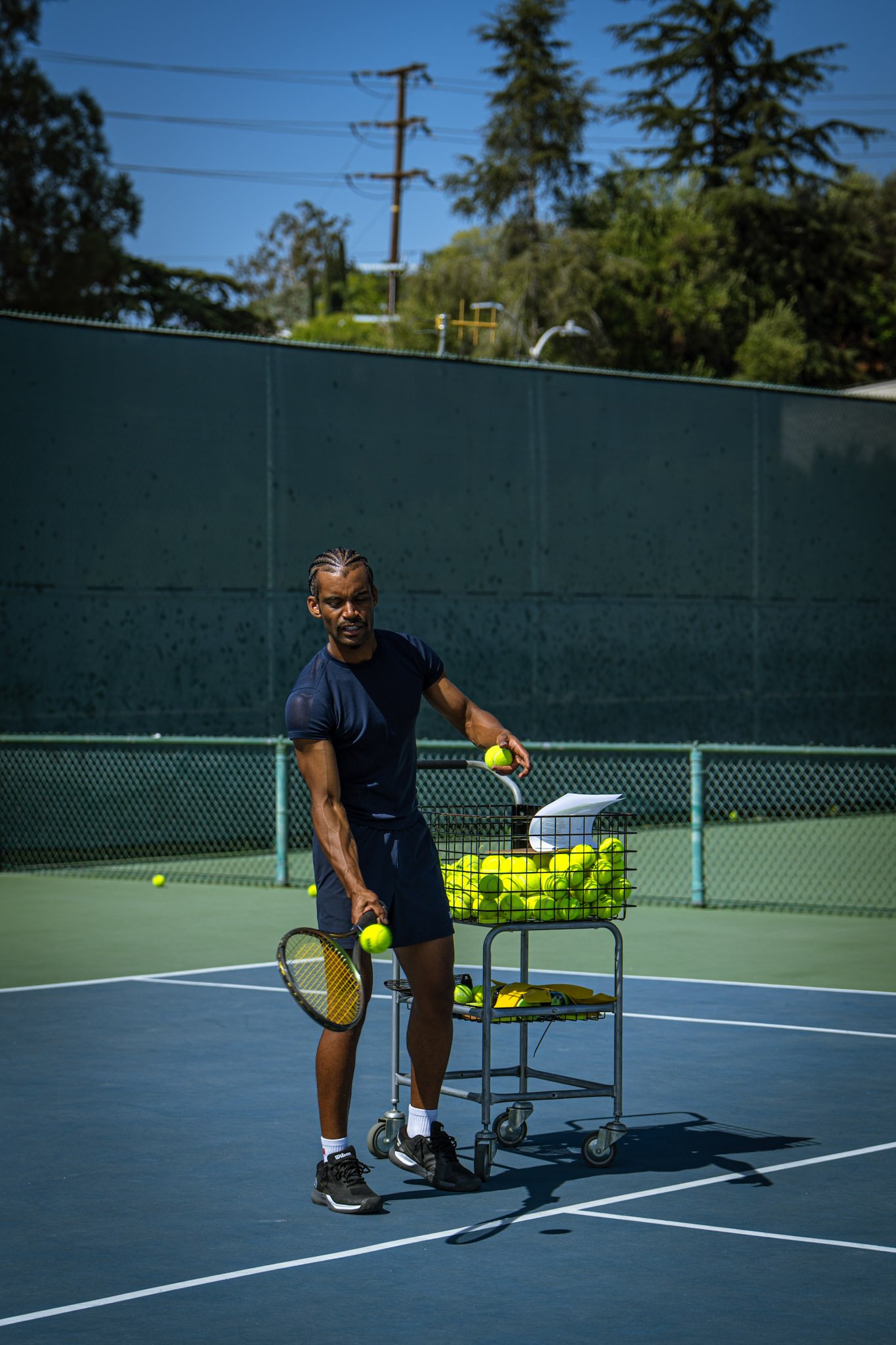 A man on a tennis court holding tennis balls and a racket, with a cart of tennis balls next to him.